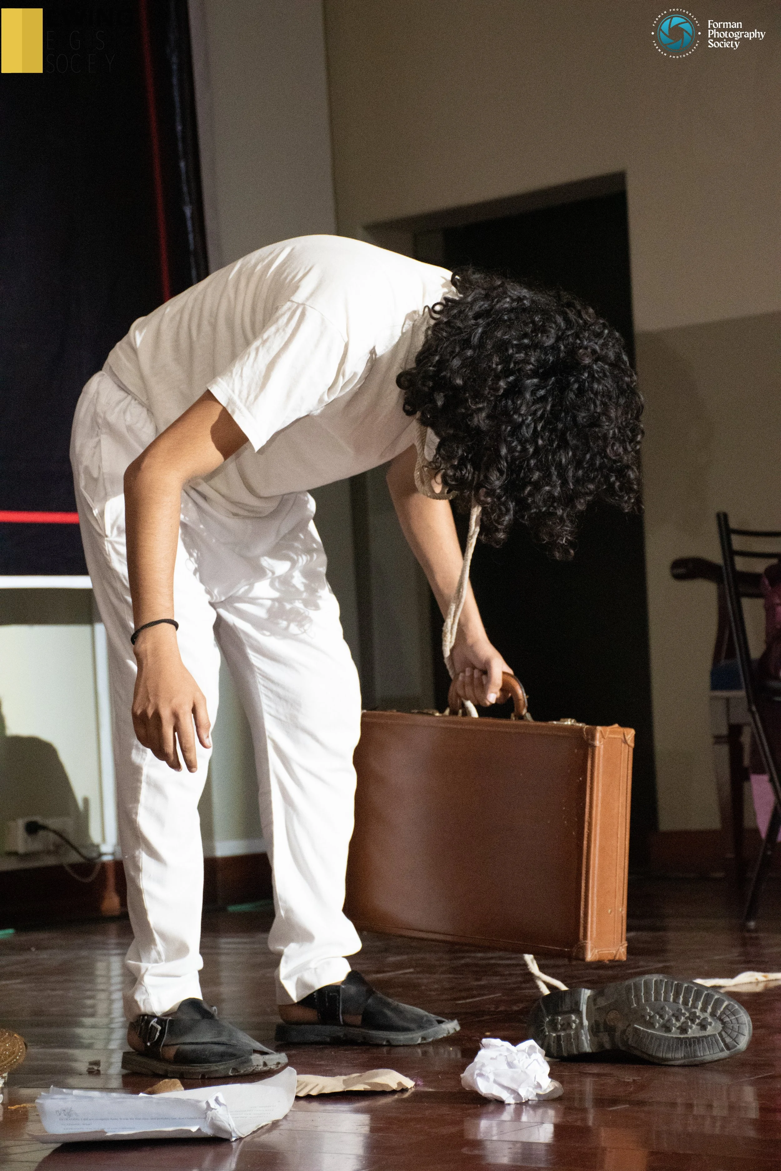Person with curly hair, dressed in white, bending down, holding a brown suitcase, on a wooden floor scattered with crumpled paper, shoes, and a paper bag, in an indoor setting.