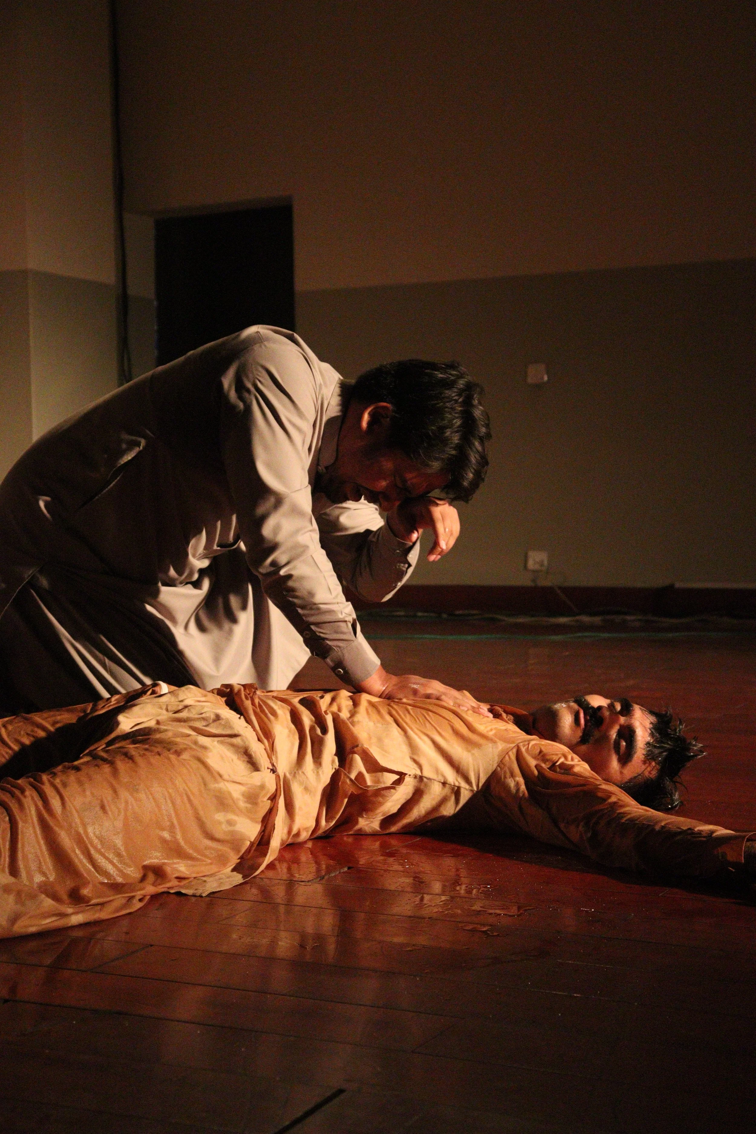 A man in a beige shirt kneels over a person lying on the floor, appearing to be mourning or distressed.