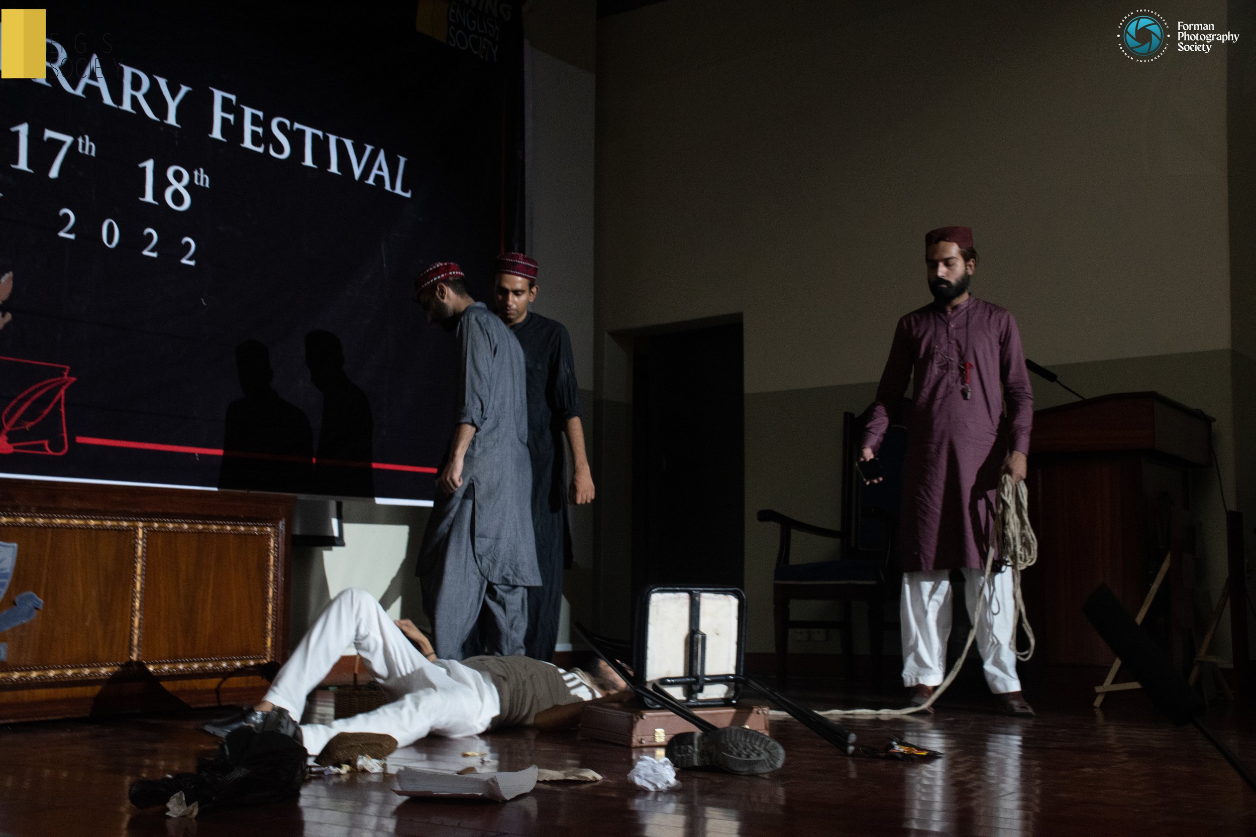 Scene from a dramatic play or reenactment at a religious festival, featuring four men in traditional clothing on a stage with props and a projected banner in the background.
