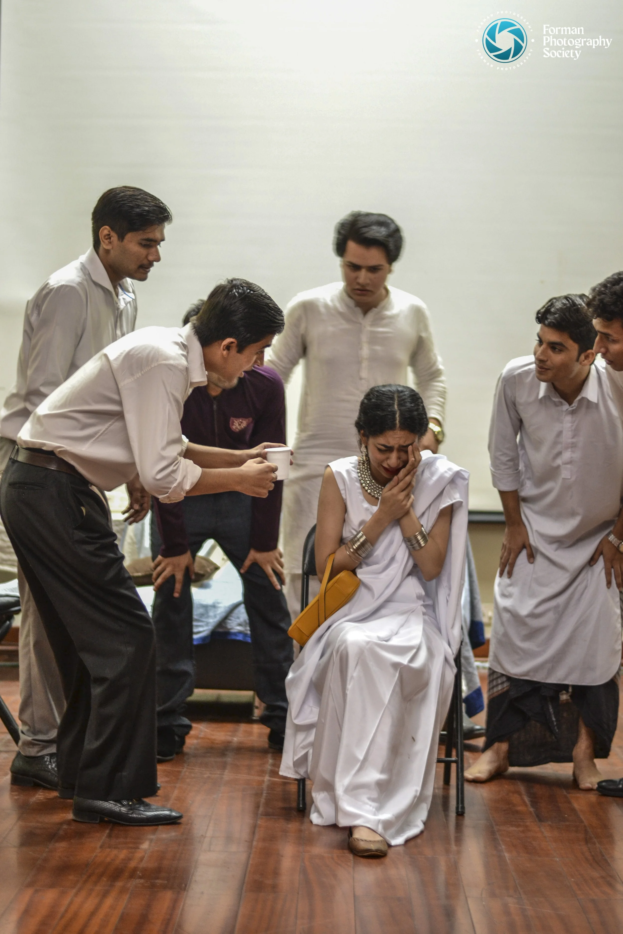 A woman dressed in white, sitting on a chair and crying, surrounded by men in traditional Indian attire who are comforting her, some with concerned expressions.