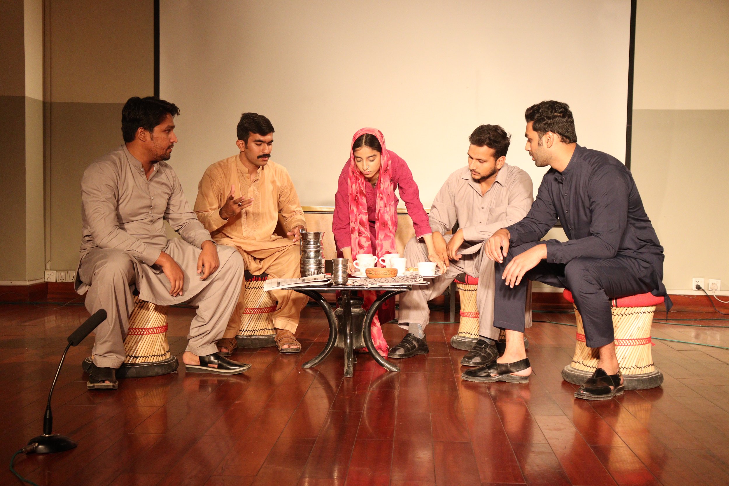 Five people sitting around a small table on stage during a theatrical performance, with one woman in a pink headscarf, engaged in a serious discussion.