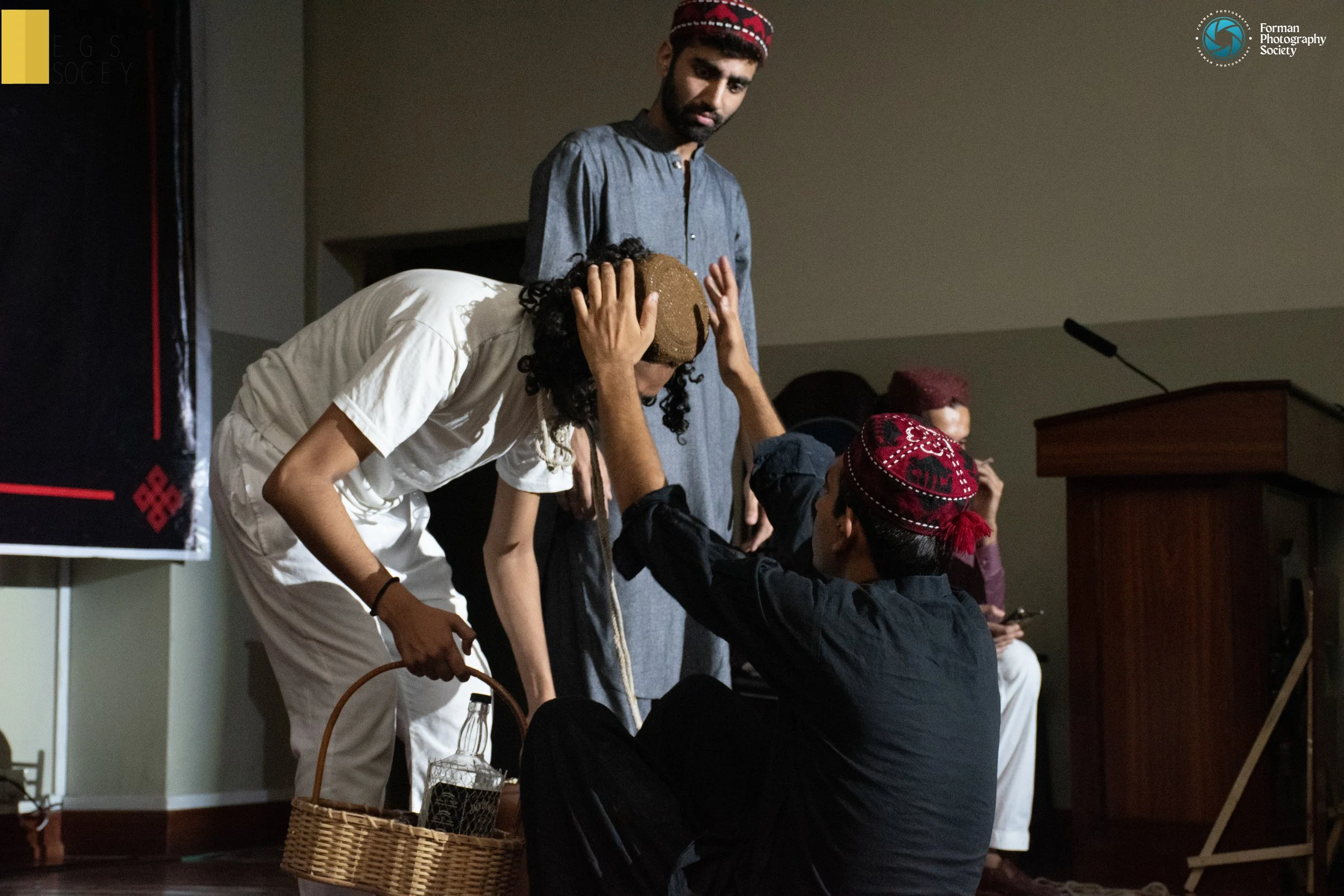 Group of people participating in a cultural or theatrical performance, with one person in black seated and wearing a red patterned cap adjusting another person's headgear, while another person in white holds a basket. A standing man observes, and a woman in the background sits near a wooden podium.