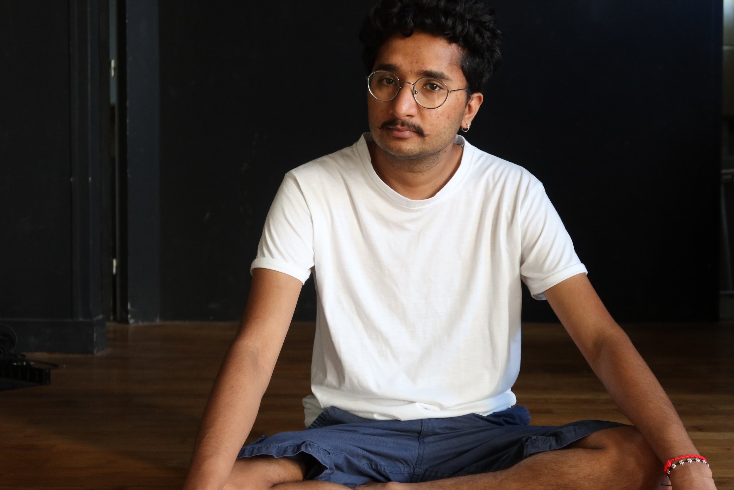 Young man with glasses, curly hair, and a mustache sitting cross-legged on wooden floor against black wall, wearing a white t-shirt and dark shorts.
