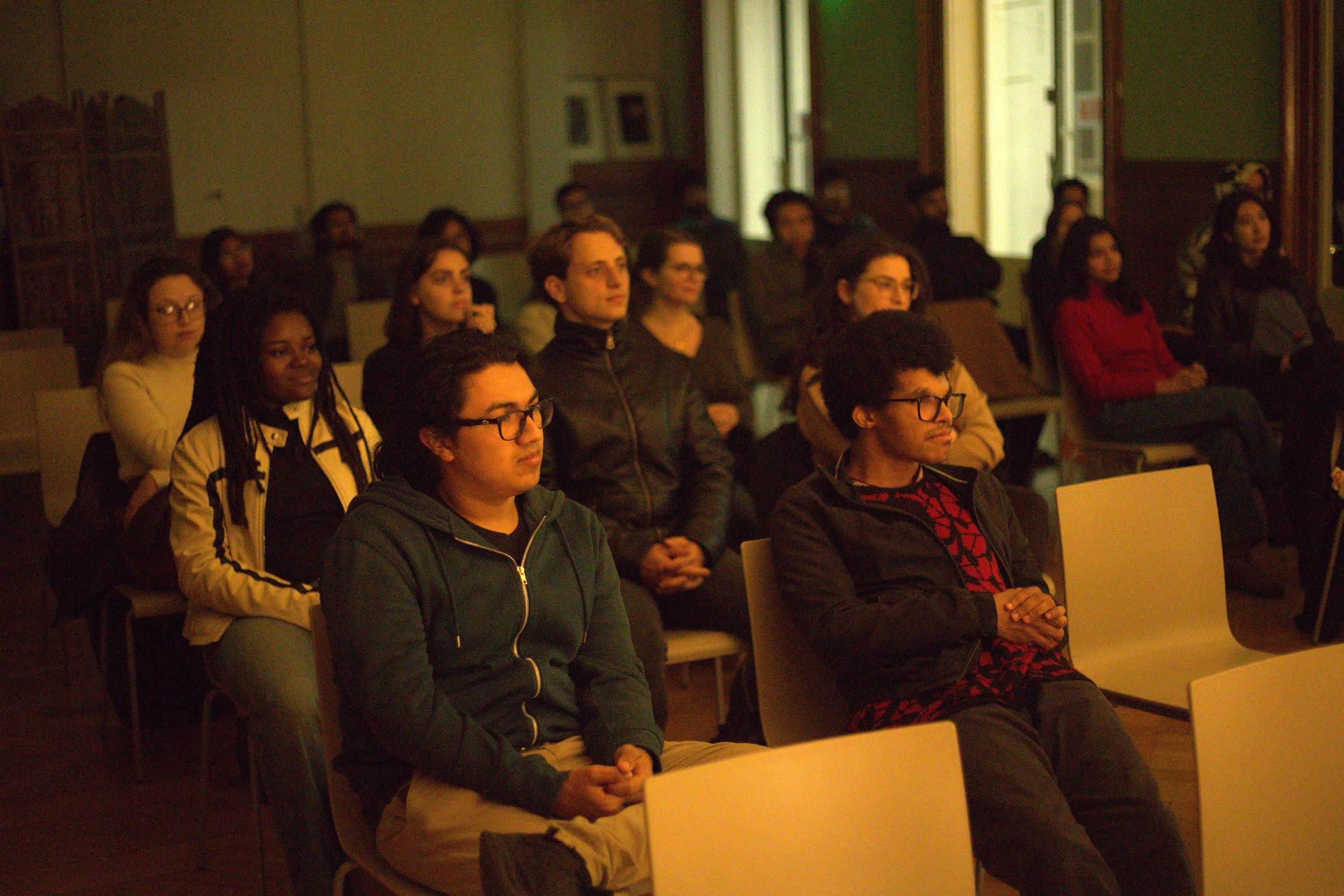 Group of people sitting attentively in a dimly lit room, possibly during a performance.