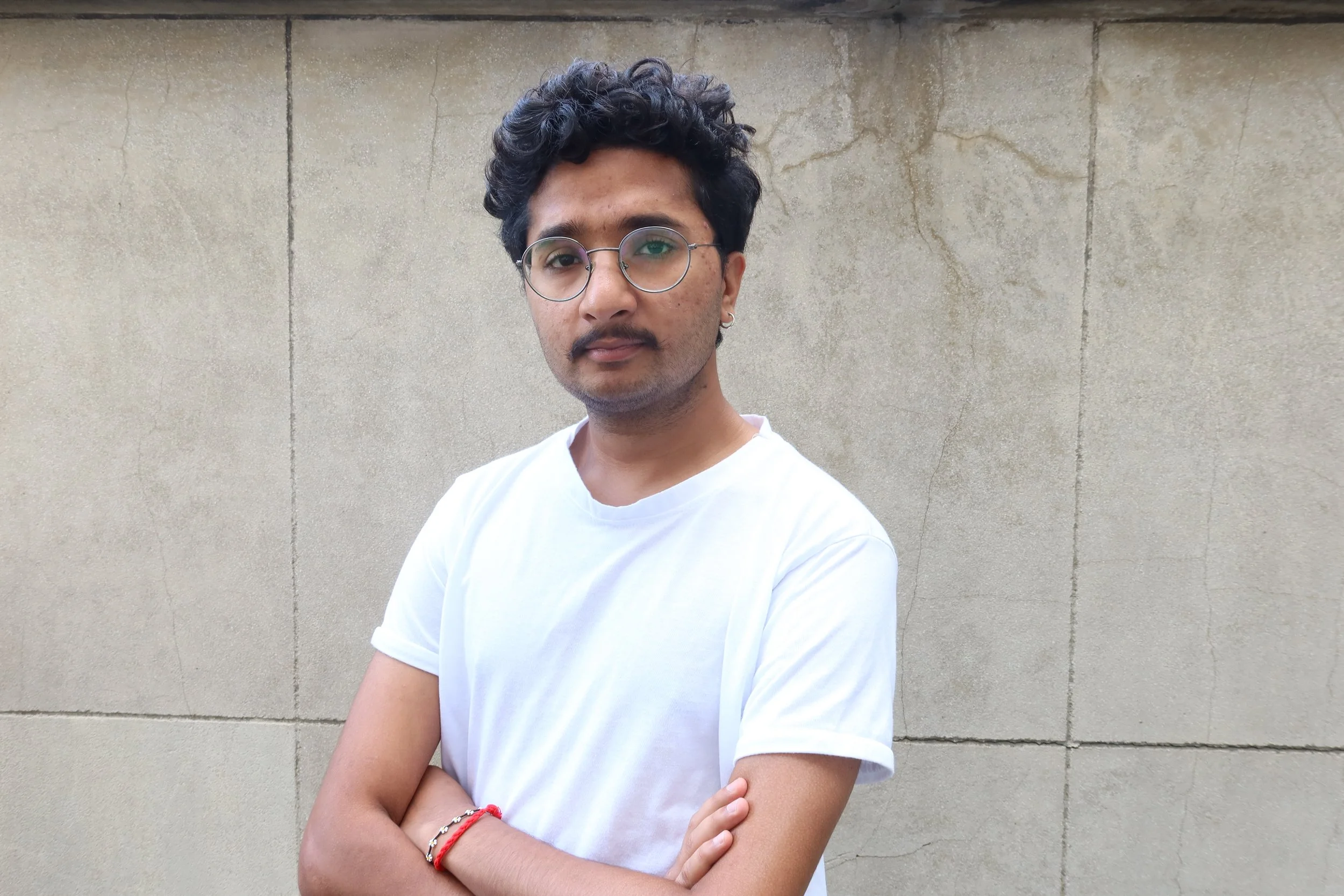 A young man with curly black hair, glasses, and earrings standing with his arms crossed in front of a plain beige wall. He is wearing a white t-shirt and a red bracelet.