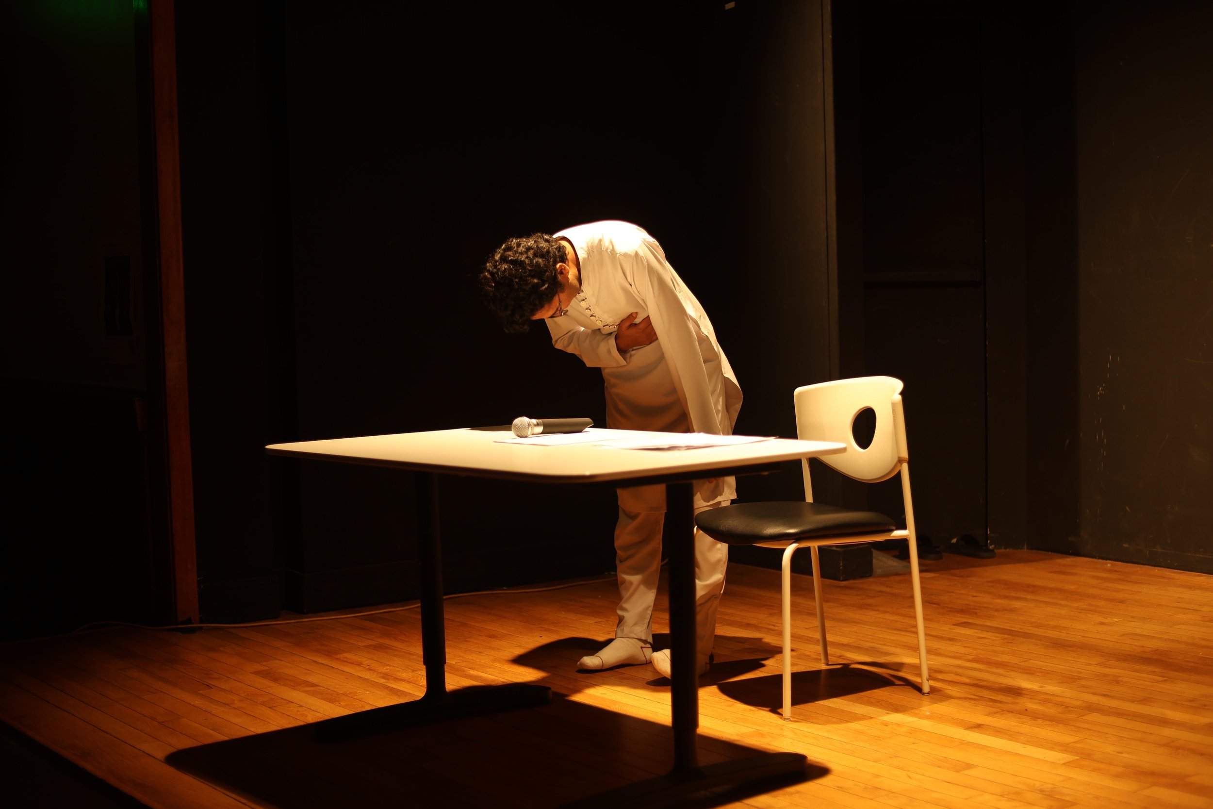 A person dressed in a white coat appears to be a doctor or scientist, bending forward and clutching their abdomen, standing behind a white table with a microphone and papers on it. There is a white chair with a black seat cushion nearby, on a wooden floor with a dark background.