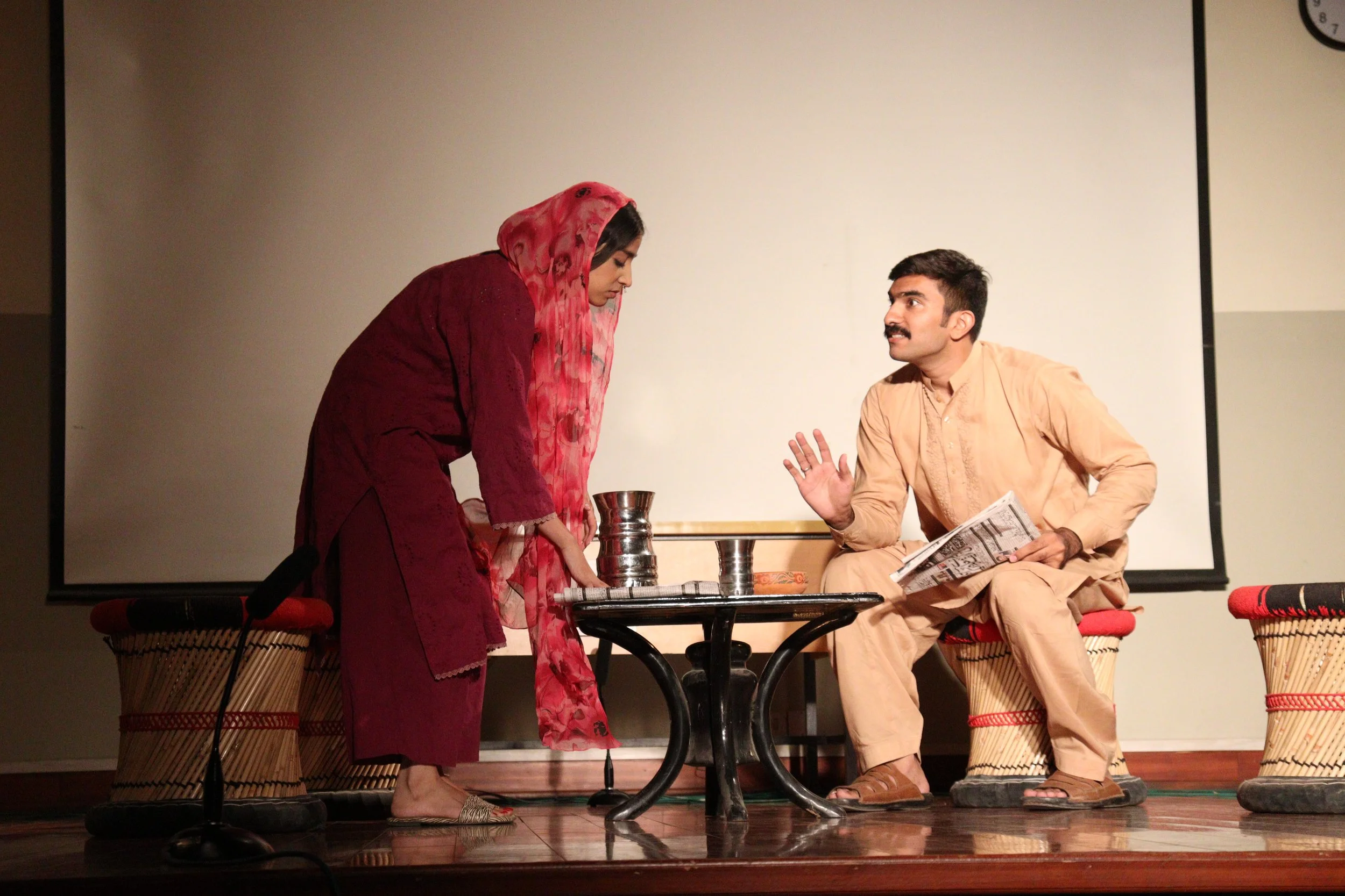 A man and a woman engaged in a conversation during a stage performance. The woman, dressed in a maroon outfit with a headscarf, is leaning over a table, while the man, in a beige traditional outfit, is sitting and gesturing with his hand. They are on a stage with a white background, surrounded by traditional Indian stools and some props on the table.