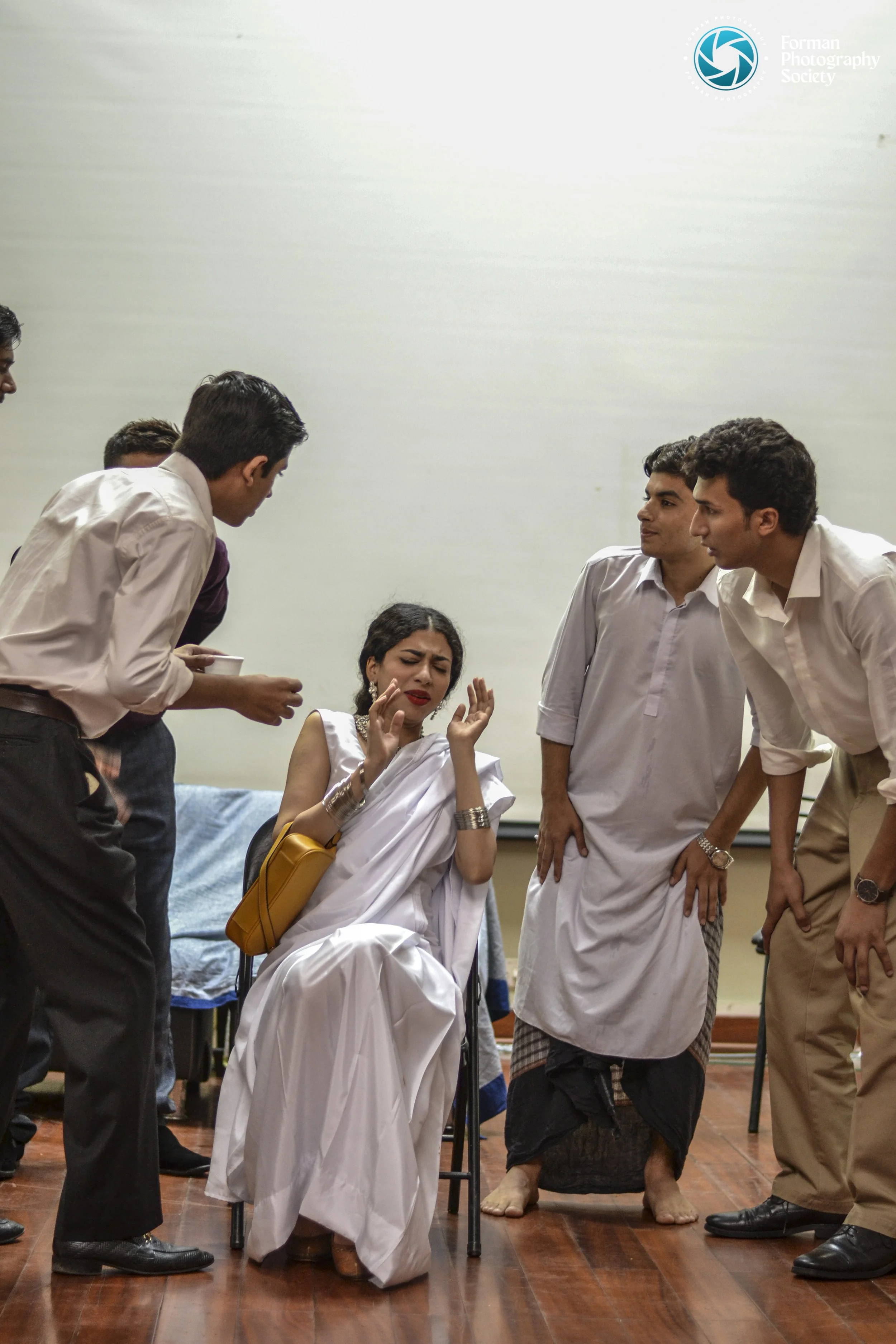 A woman in a white saree with a yellow bag appears to be upset or in distress, surrounded by several men in traditional or formal attire, some bowing their heads or leaning forward, in an indoor setting with a wooden floor.