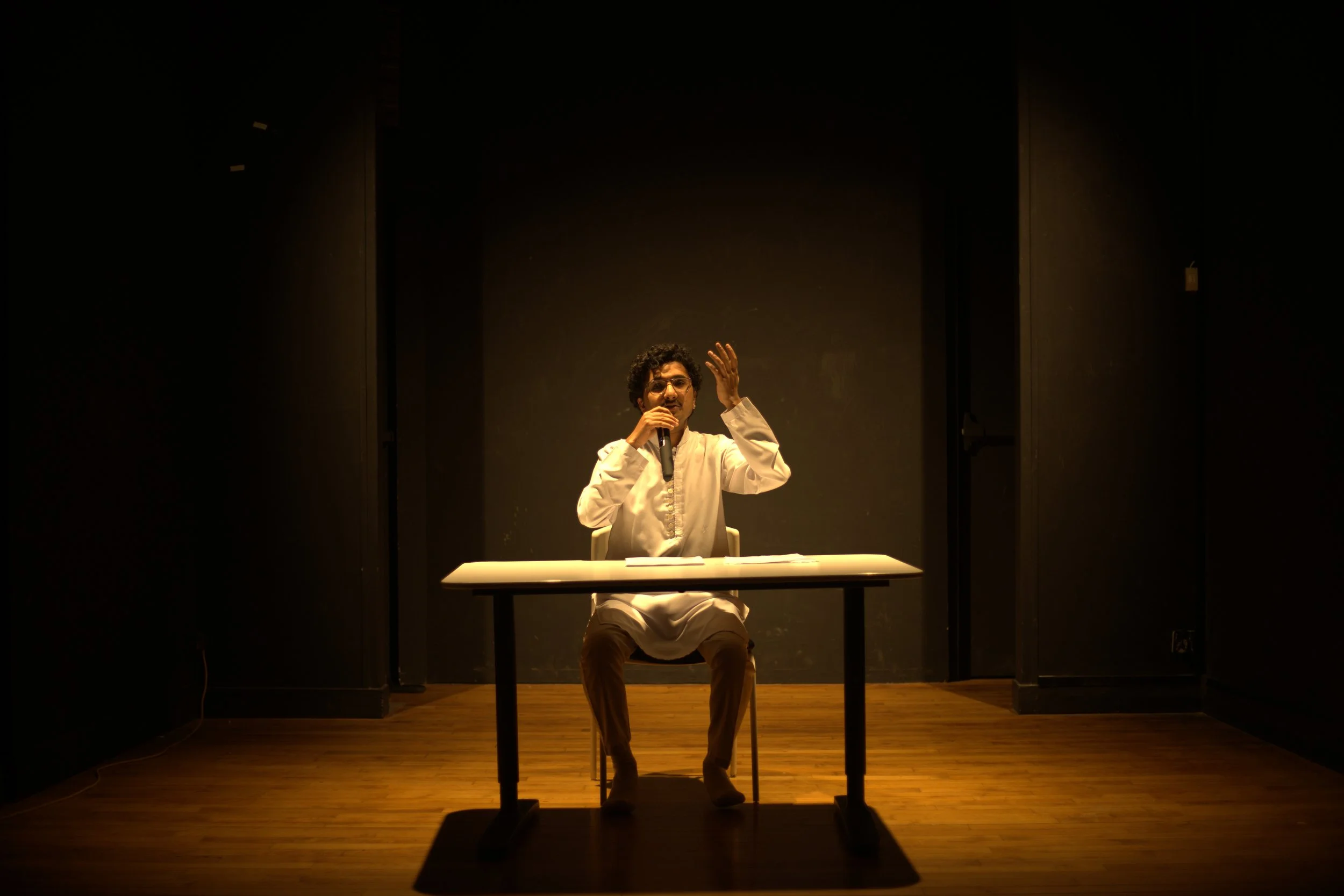 Person sitting at a table in a dimly lit room, speaking into a microphone, with a dark background and wooden floor.