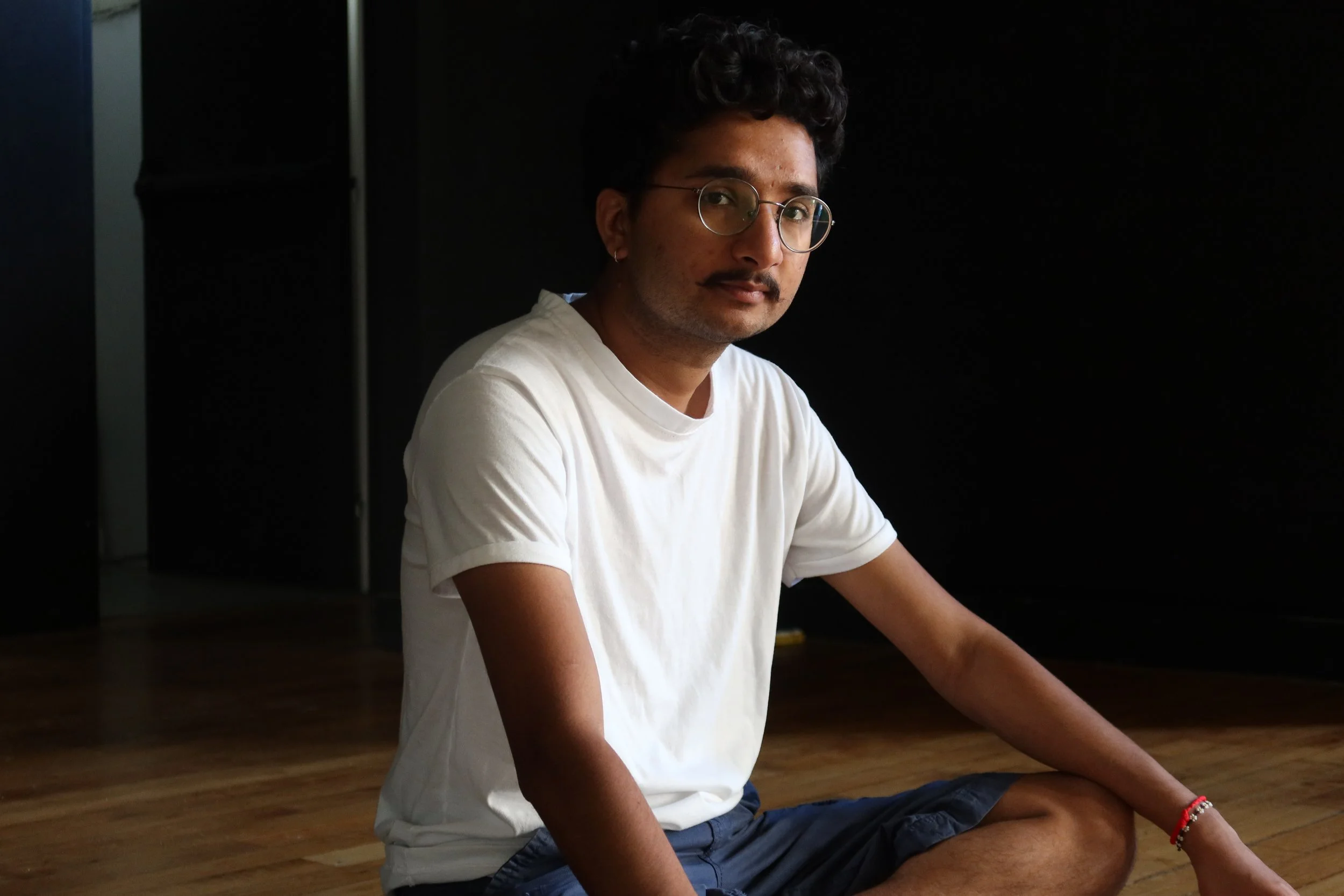 A young man with dark curly hair, glasses, and a mustache, wearing a white t-shirt and shorts, sitting on a wooden floor against a dark background.
