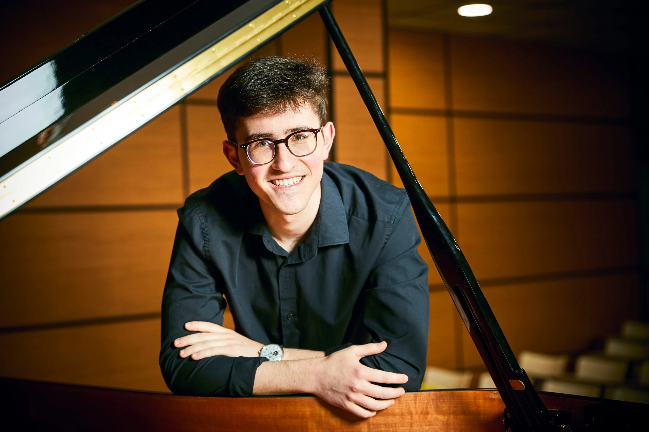 Joven con gafas y camisa negra, apoyado en un piano de cola, en una sala de conciertos con paredes de madera.