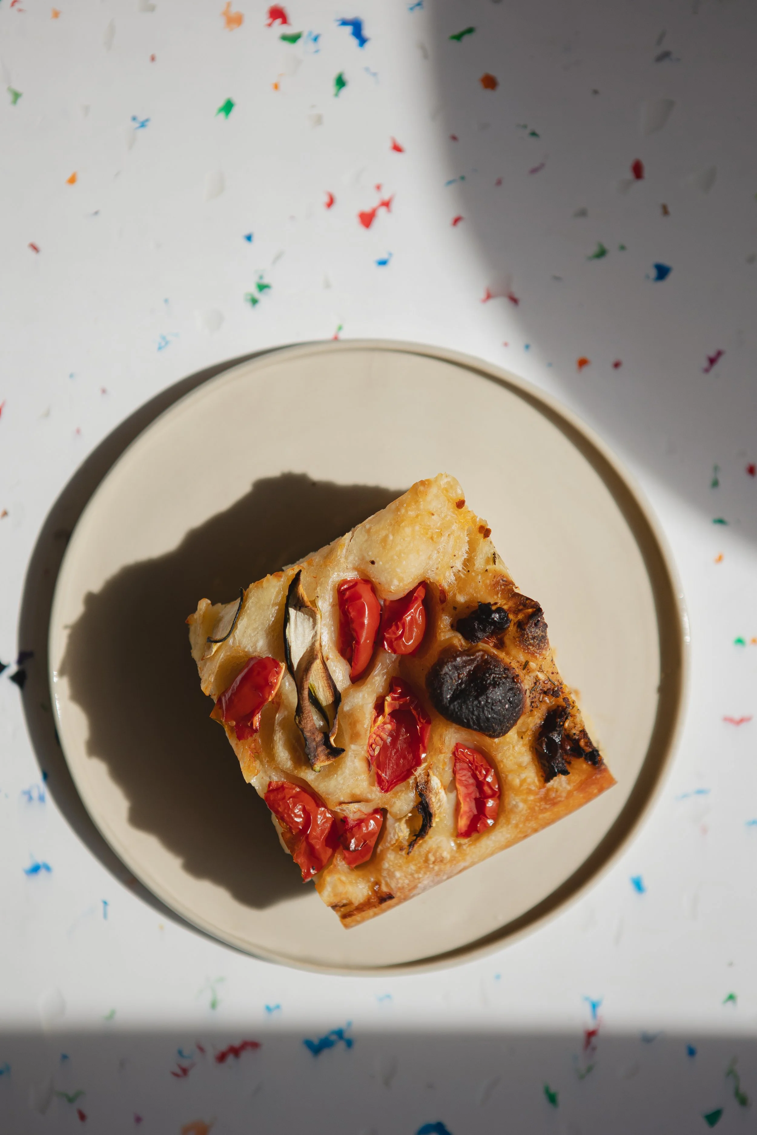 Ein Stück Pizza mit Tomaten, Käse und schwarzen Oliven auf einem weißen Teller, bei Sonnenlicht, bunter Konfetti im Hintergrund.