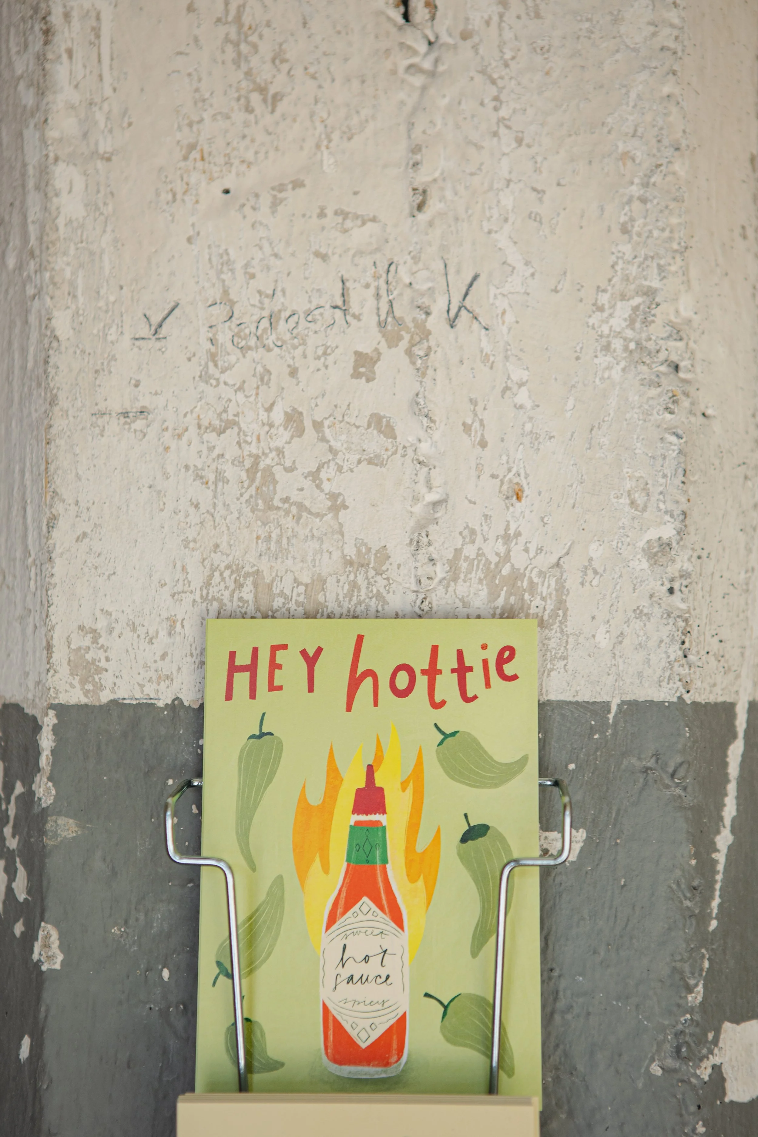 Ein handgemaltes Schild mit der Aufschrift "HEY hottie" und einem Guajak-Fleisch, umgeben von Paprika, an einem grauen, alten Wand. Über dem Schild ist eine wiederverwenden, mit Bleistift geschriebene Notiz auf der Wand.