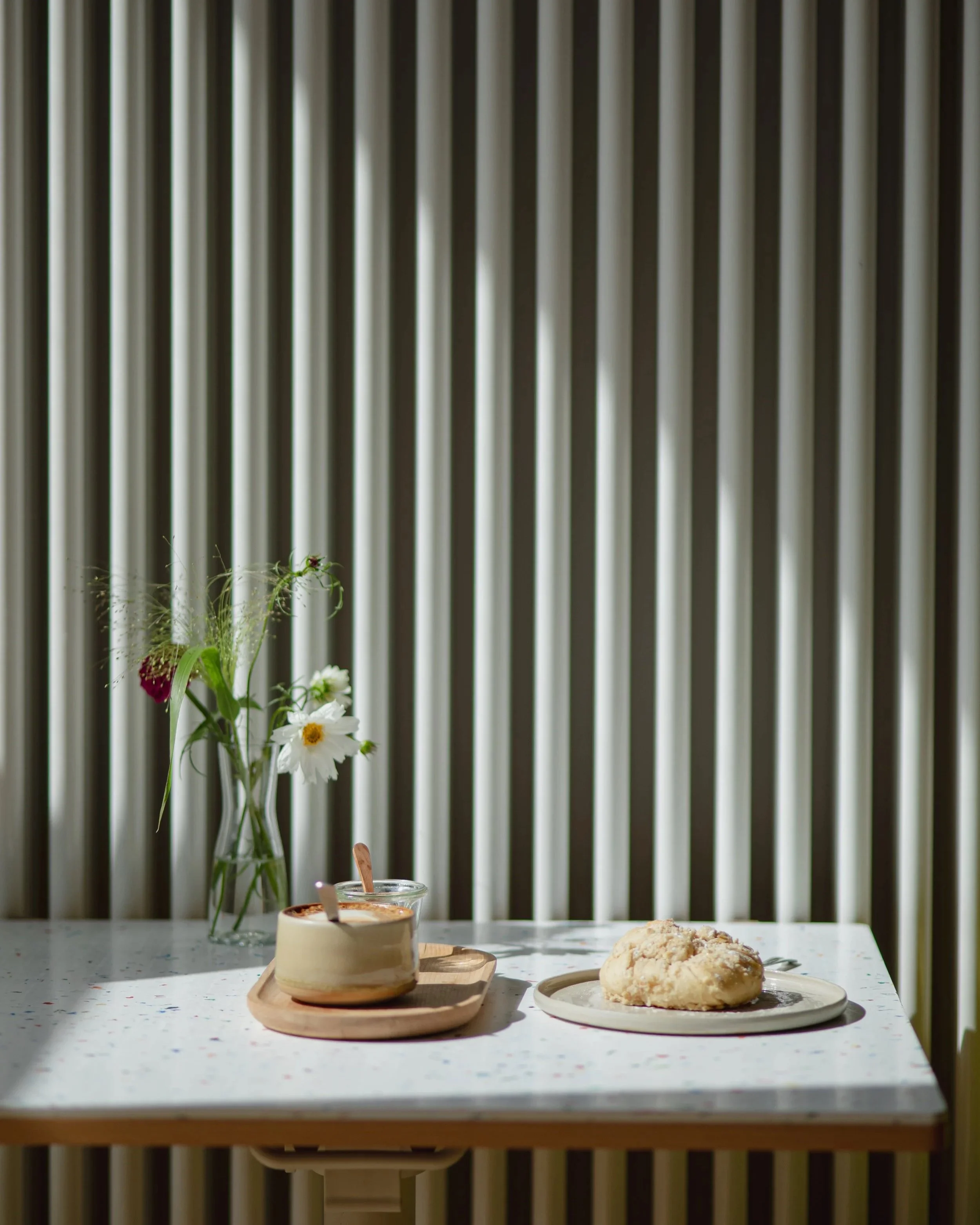 Ein Tisch mit einer Vase mit Blumen, einem Becher, einem Löffel, und einem Gebäckstück in einem Raum mit vertikalen Holzlamellen-Wänden.