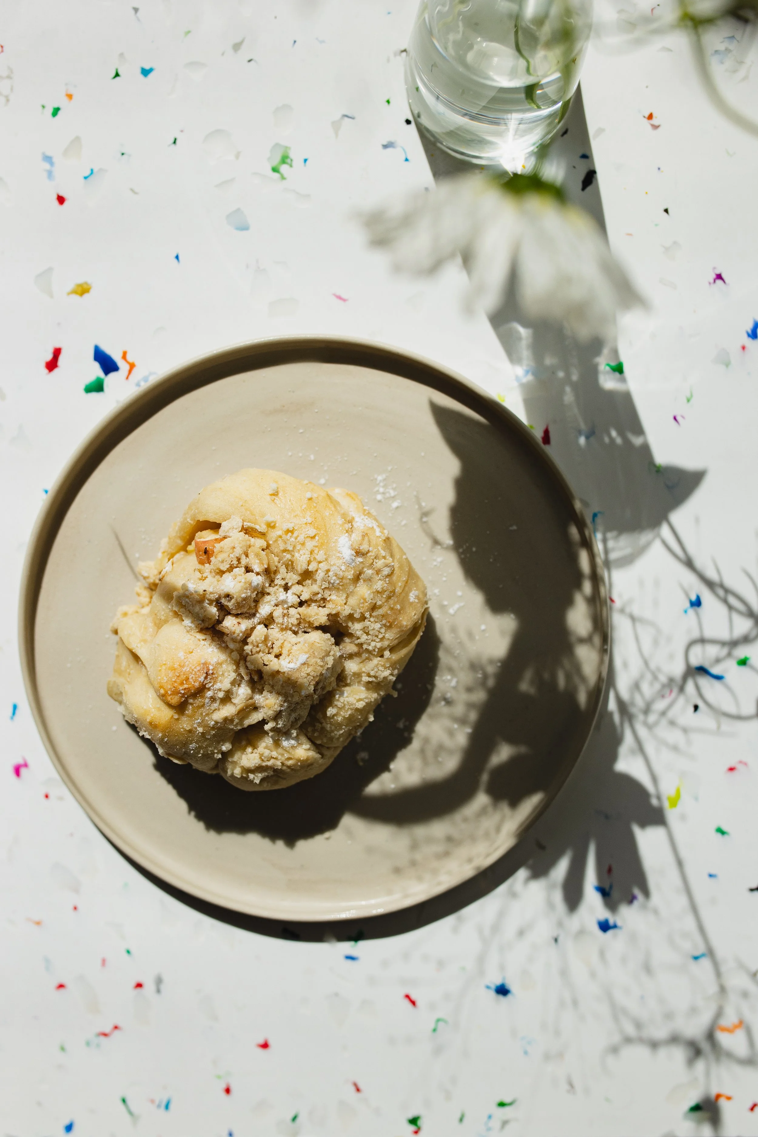 Zimtschnecke  auf einem selbst getöpferten Teller weißen Tisch, dekoriert mit buntem Konfetti, neben einem Glas Wasser. Schatten einer Blumenranke ist sichtbar.