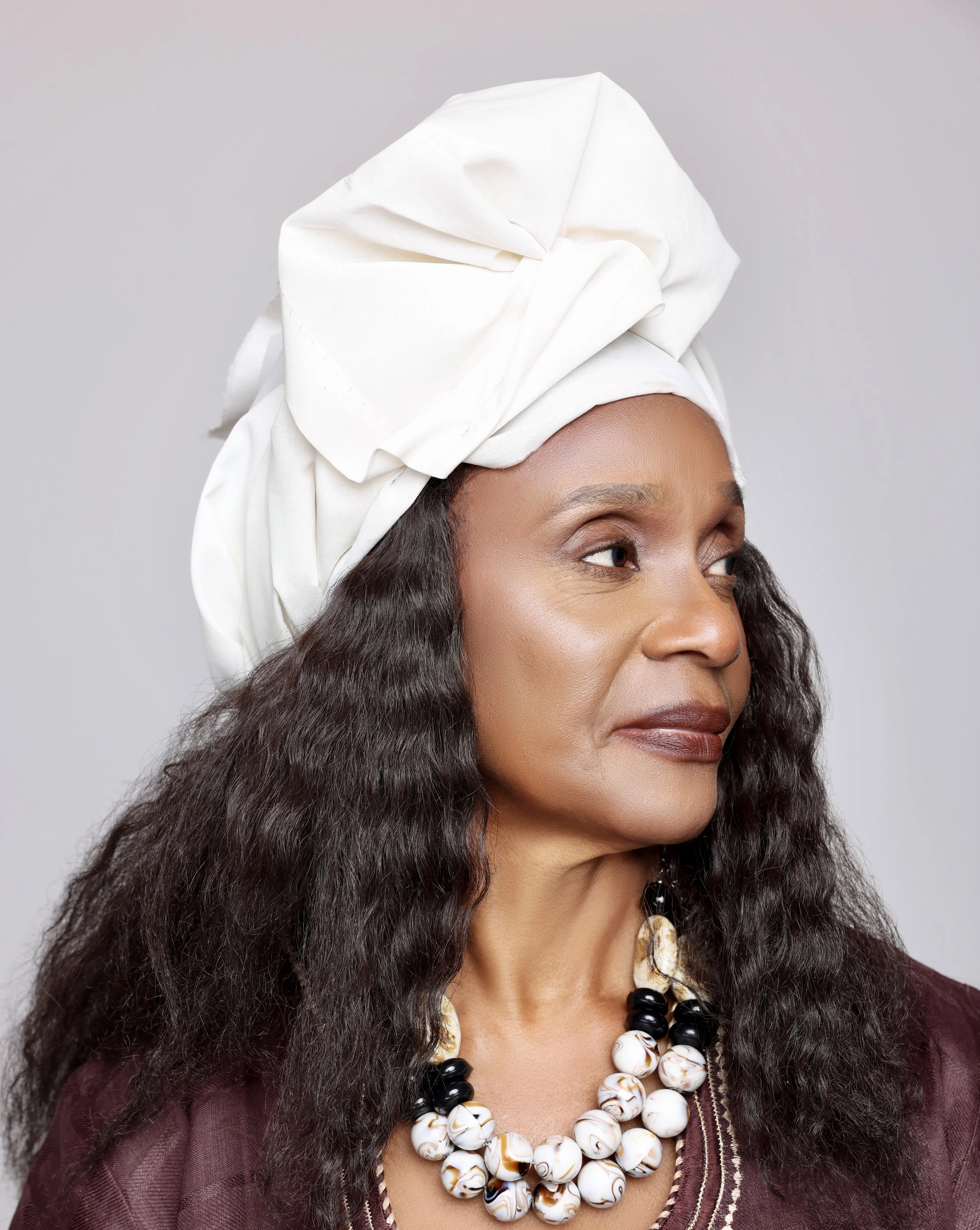 A woman with dark curly hair wearing a large white headwrap, a beaded necklace, and a maroon top, looking to her right with a neutral expression.