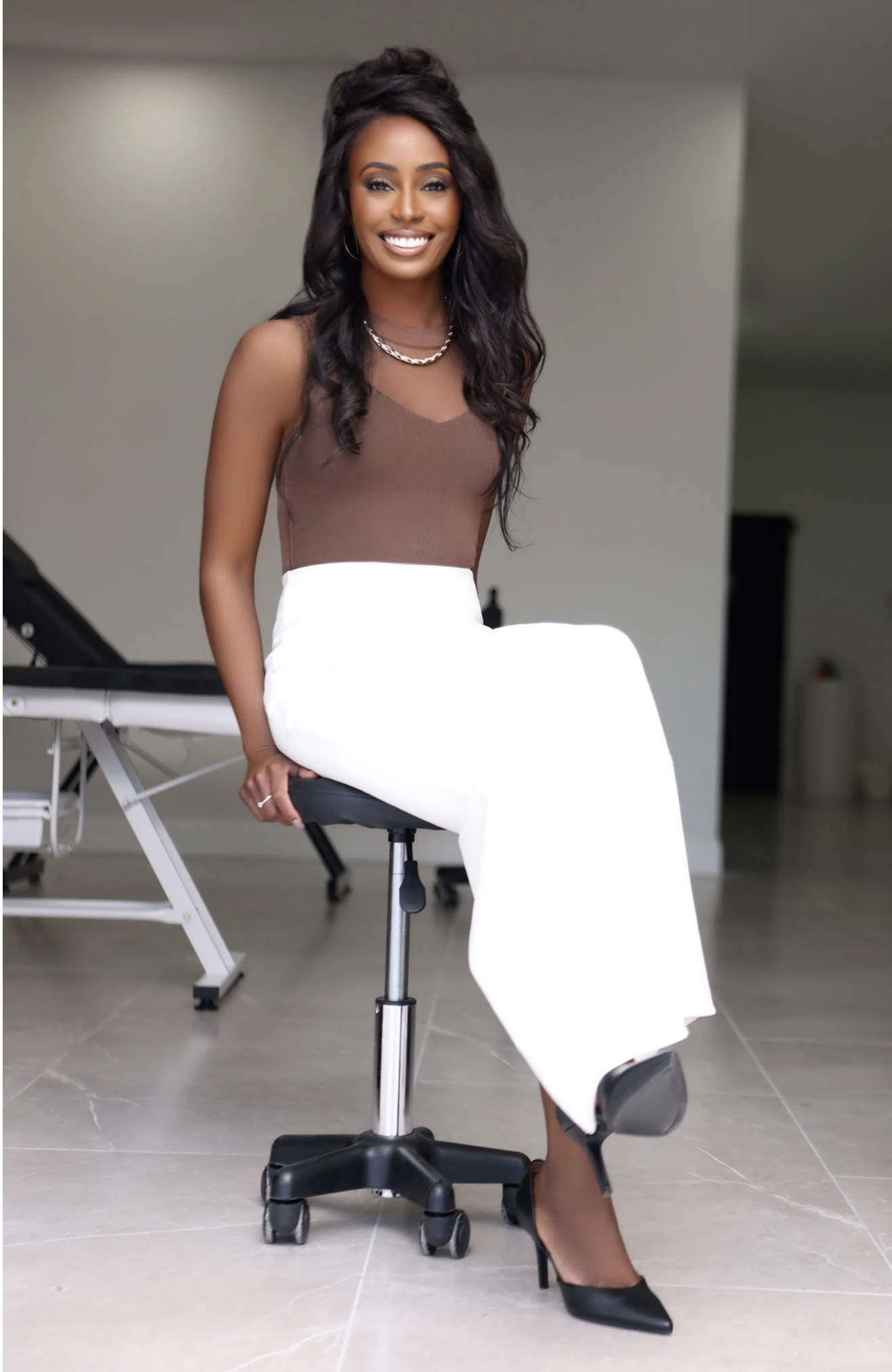 A woman with long dark hair, wearing a brown sleeveless top, white wide-leg pants, and black heels, sitting on an office chair in a modern indoor space, smiling at the camera.