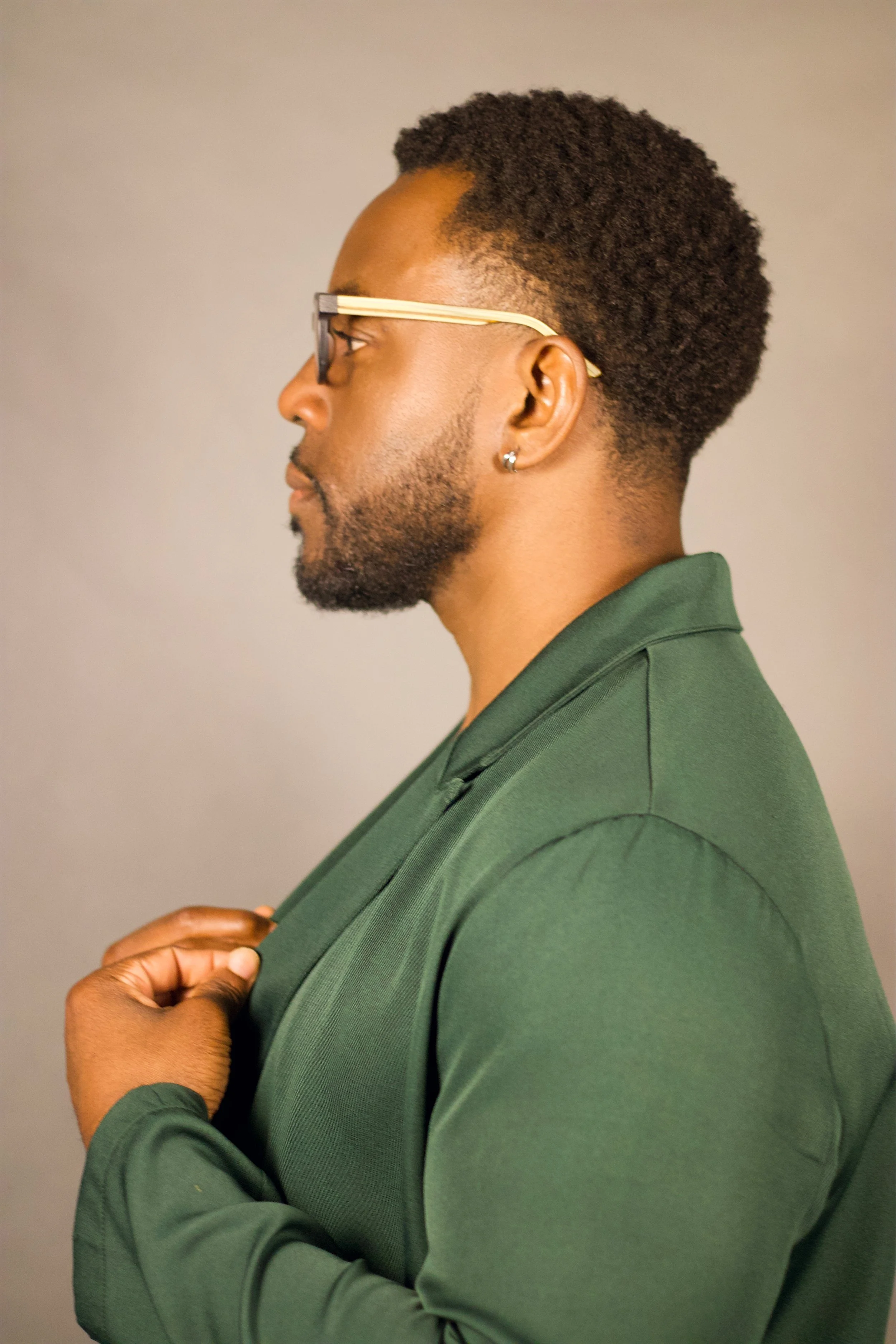 Side profile of a man with glasses, earring, short textured hair, wearing a dark green collared shirt, against a neutral background.