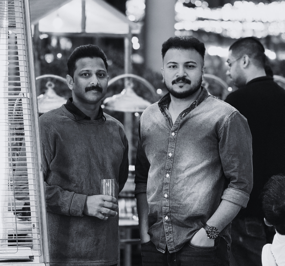 Two men standing together at a social gathering, one holding a glass, with other people in the background.