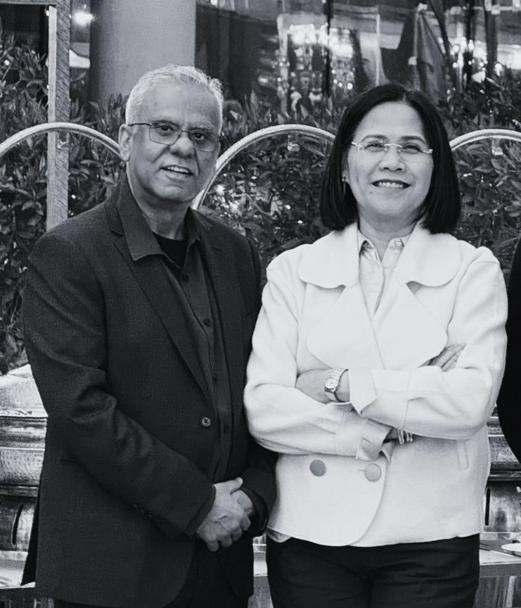 Two adults, a man and a woman, standing side by side and smiling in an indoor location with plants and decorative lighting in the background.