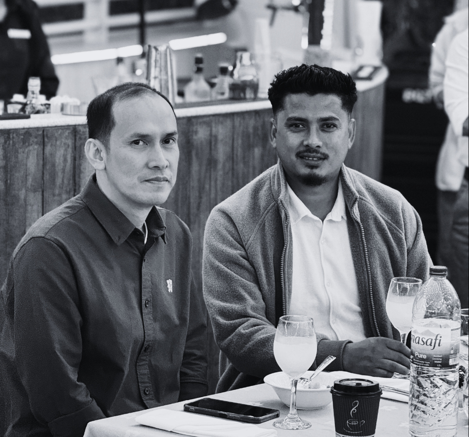 Two men sitting at a restaurant table, with drinks and a smartphone in front of them. One man has short hair and is wearing a button-up shirt, while the other has a styled haircut and is wearing a jacket over a white shirt.