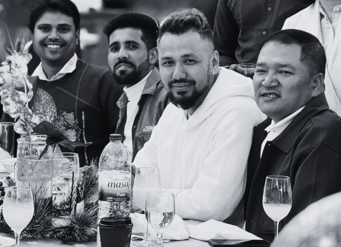 Group of five men sitting at a table with drinks and a floral centerpiece, smiling at an indoor gathering.