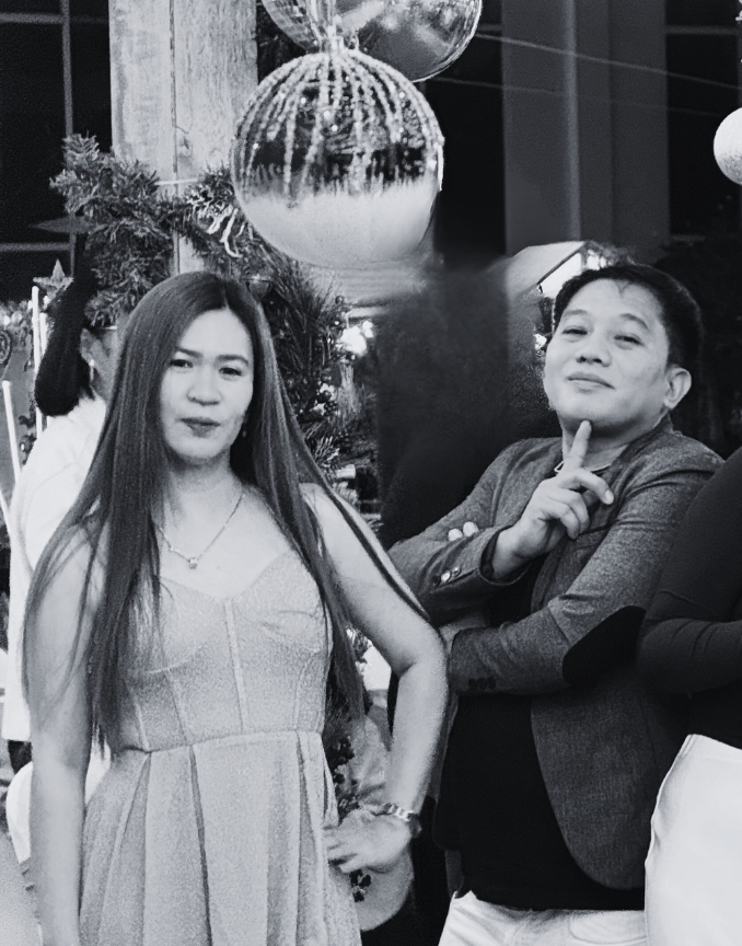 Two people posing in front of a decorated Christmas tree with large ornaments, one woman with long hair and a dress, and a man with short hair and a jacket making a finger gun gesture