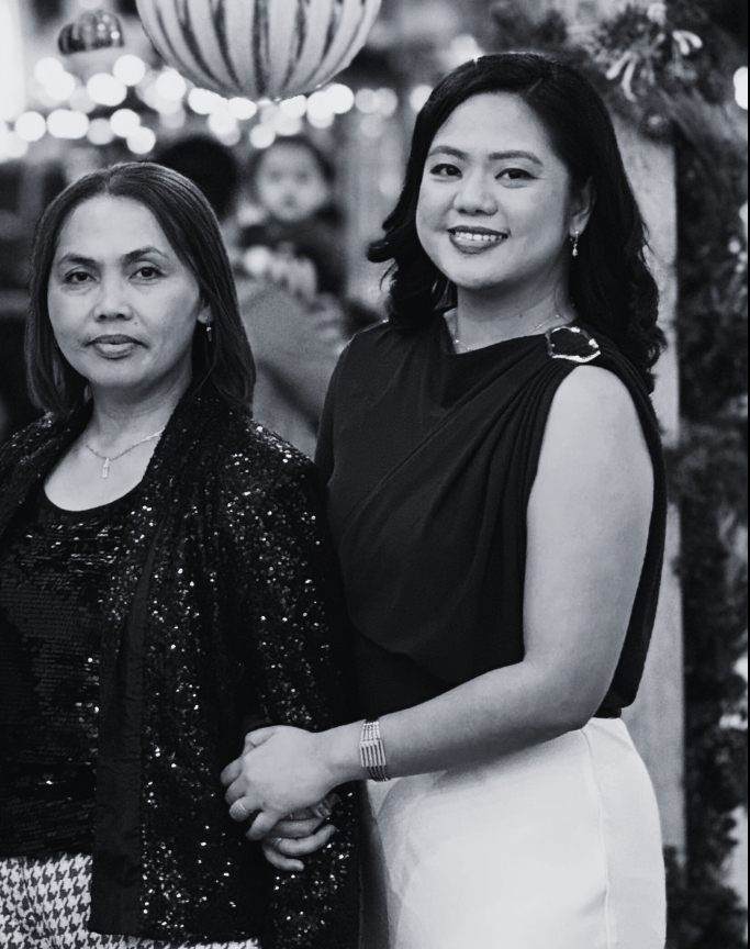 Two women standing close together at a festive event, smiling at the camera. The woman on the left has shoulder-length hair, wearing a sparkly black jacket and black top. The woman on the right has medium-length hair, wearing a sleeveless black top a
