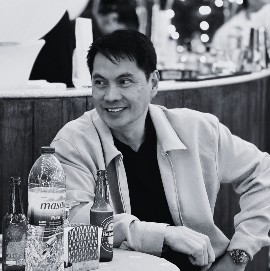 A man seated at a table, smiling and looking to his left in a restaurant or social setting with bottles of soda and beer on the table.