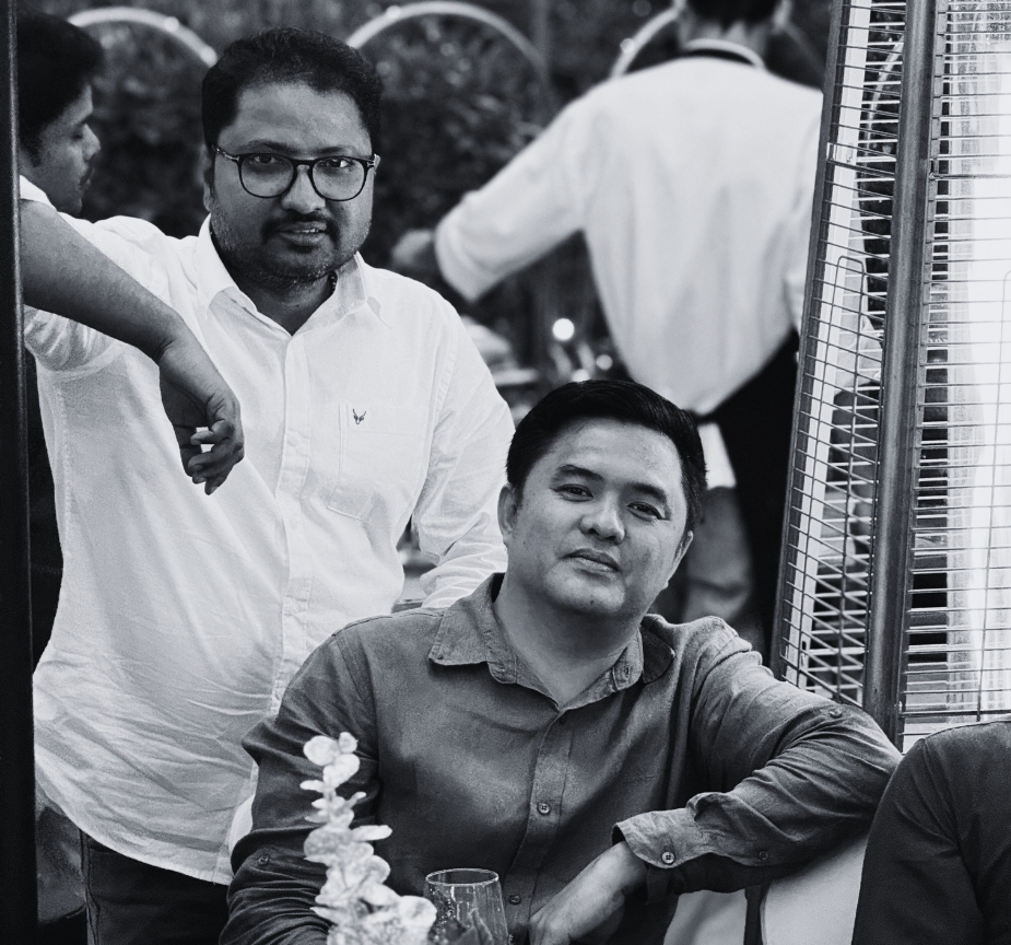 Two men at a social gathering, one with glasses standing and another sitting beside a table, in front of a tall outdoor heater.