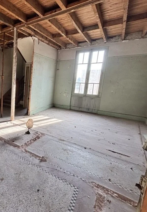 Une pièce en cours de rénovation avec un sol partiellement couvert de tapis ou de matériaux de construction, un mur avec une fenêtre laissant entrer la lumière naturelle, et une partie du plafond en bois non fini.