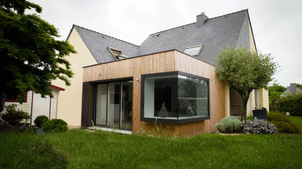 Une maison moderne avec une façade en bois et de grandes fenêtres, entourée d'un jardin verdoyant avec des arbustes et des arbres.