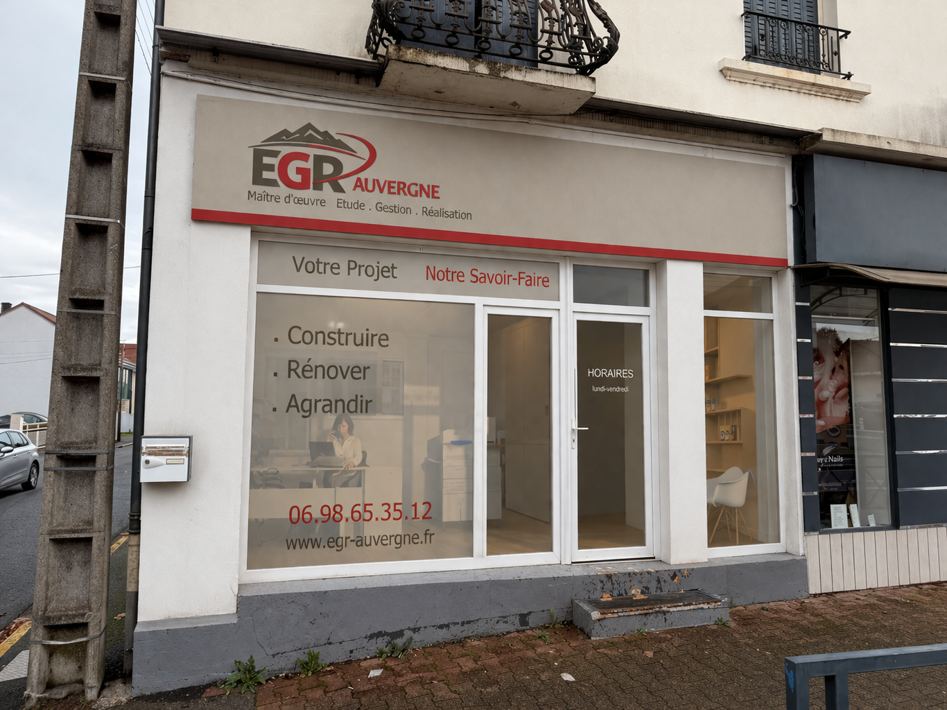 Vitrine d'une agence  avec le nom 'EGR AUVERGNE'. La vitrine indique des services de construction, rénovation, agrandissement, et affiche un numéro de téléphone et un site web. La porte donne accès à un espace intérieur avec une personne assise à un bureau.