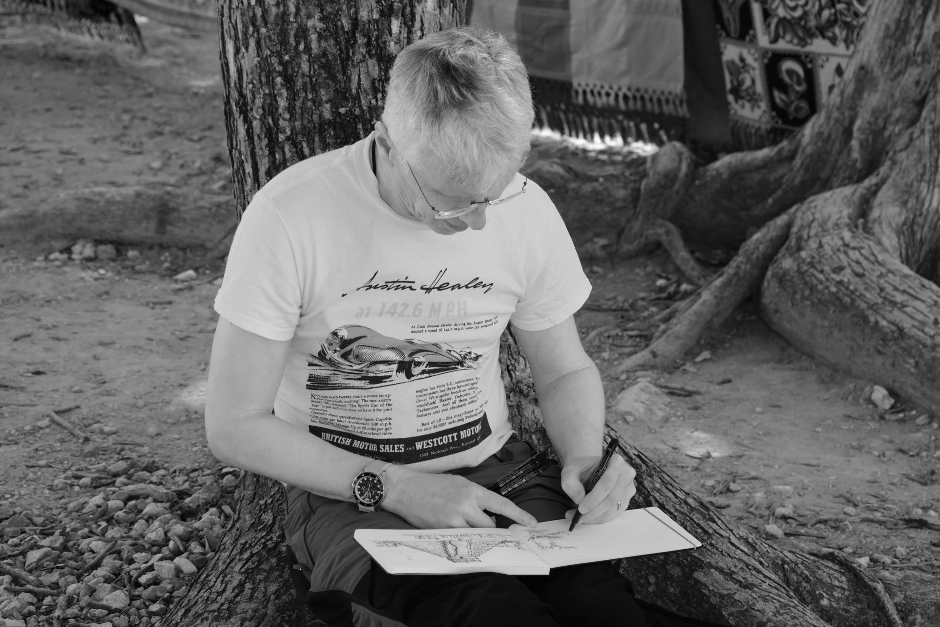 A man with light-colored hair, glasses, and a watch, sitting outdoors against a tree, writing or sketching in a notebook on his lap.