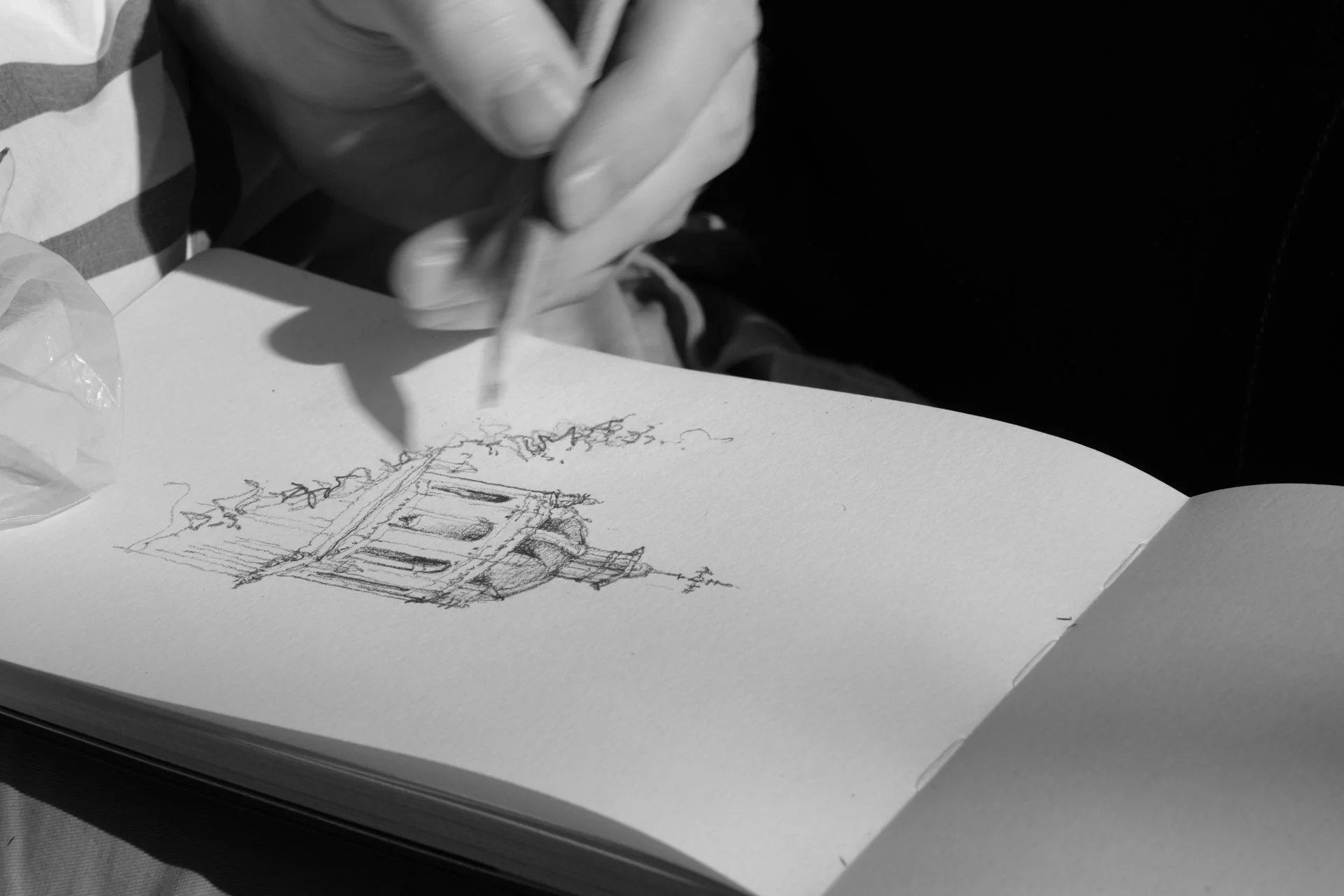 A person wearing a striped shirt and gloves is sketching a building with intricate details on a piece of paper, with a container of ink nearby.