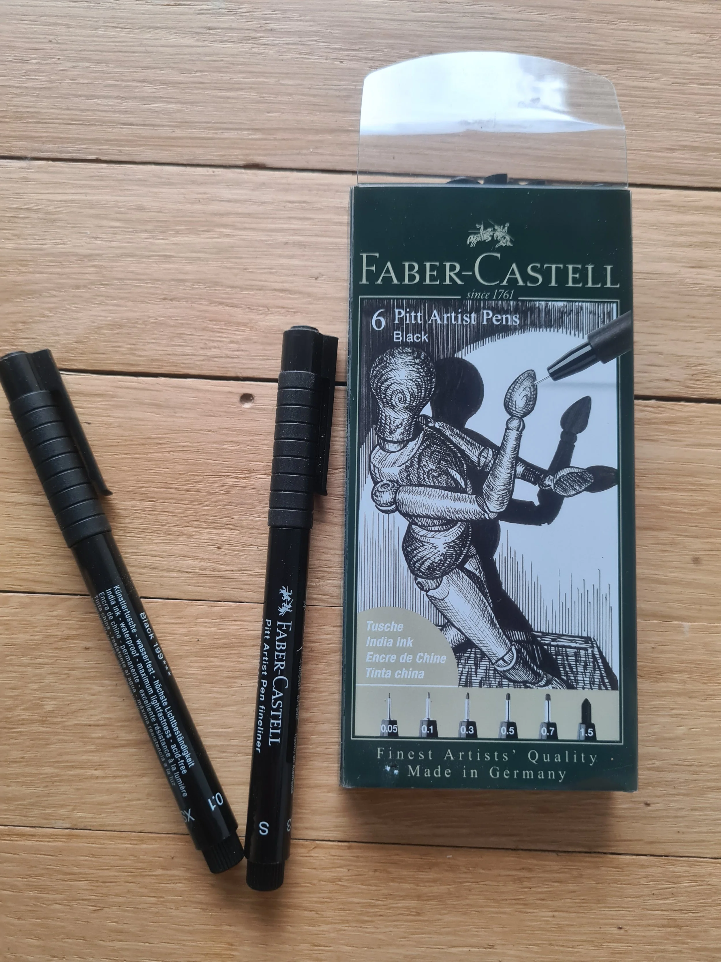 Box of Faber-Castell Pitt Artist Pens in black, with two pens placed beside it on a wooden surface.
