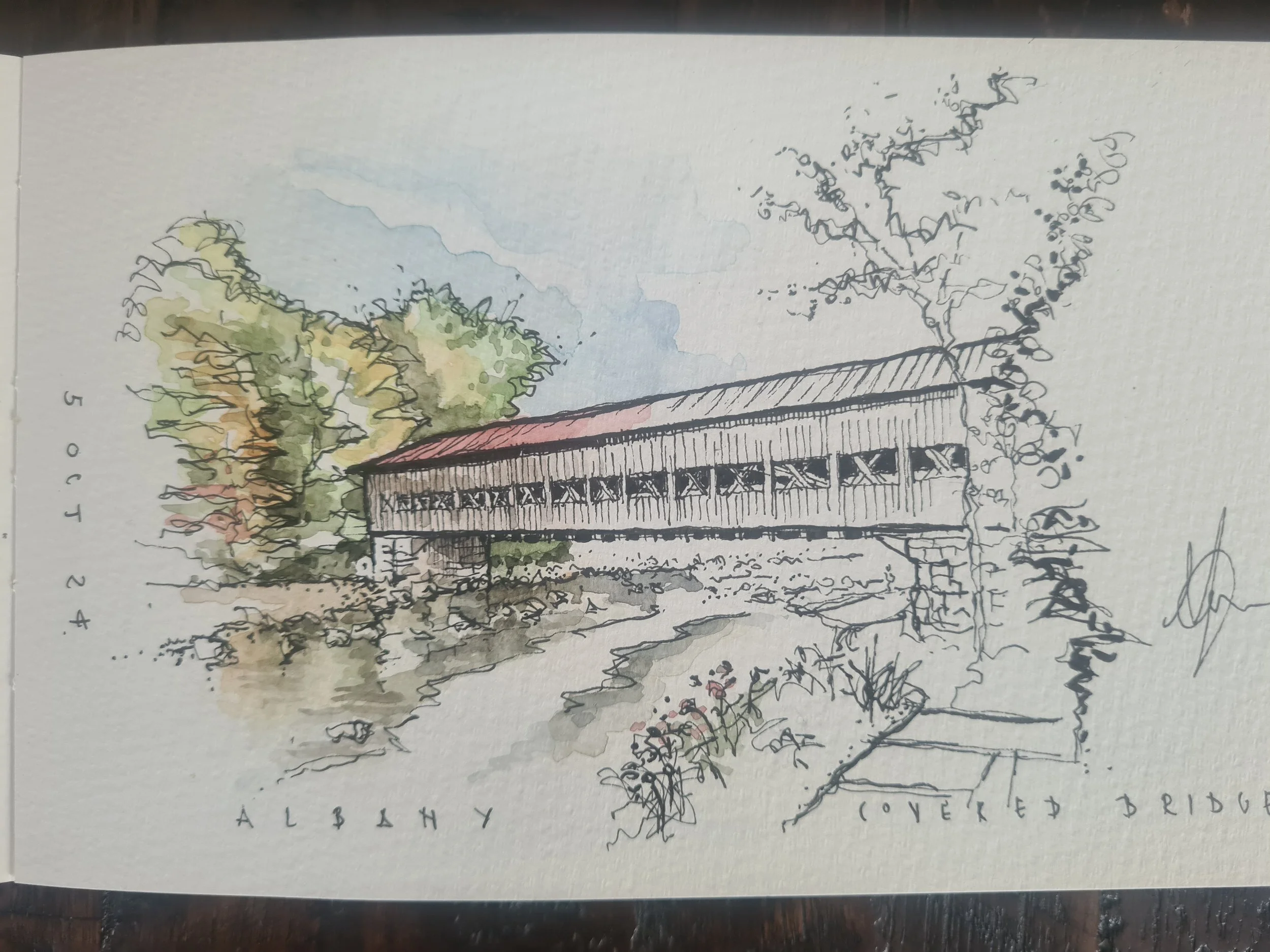 A watercolor sketch of a covered bridge in an alfalfa field with trees in the background. The bridge is raised on stone supports and has a red roof, with the words 'Covered Bridge' written on it. The words 'Alfalfa' and '5 OCT 24' are also visible on the sketch.
