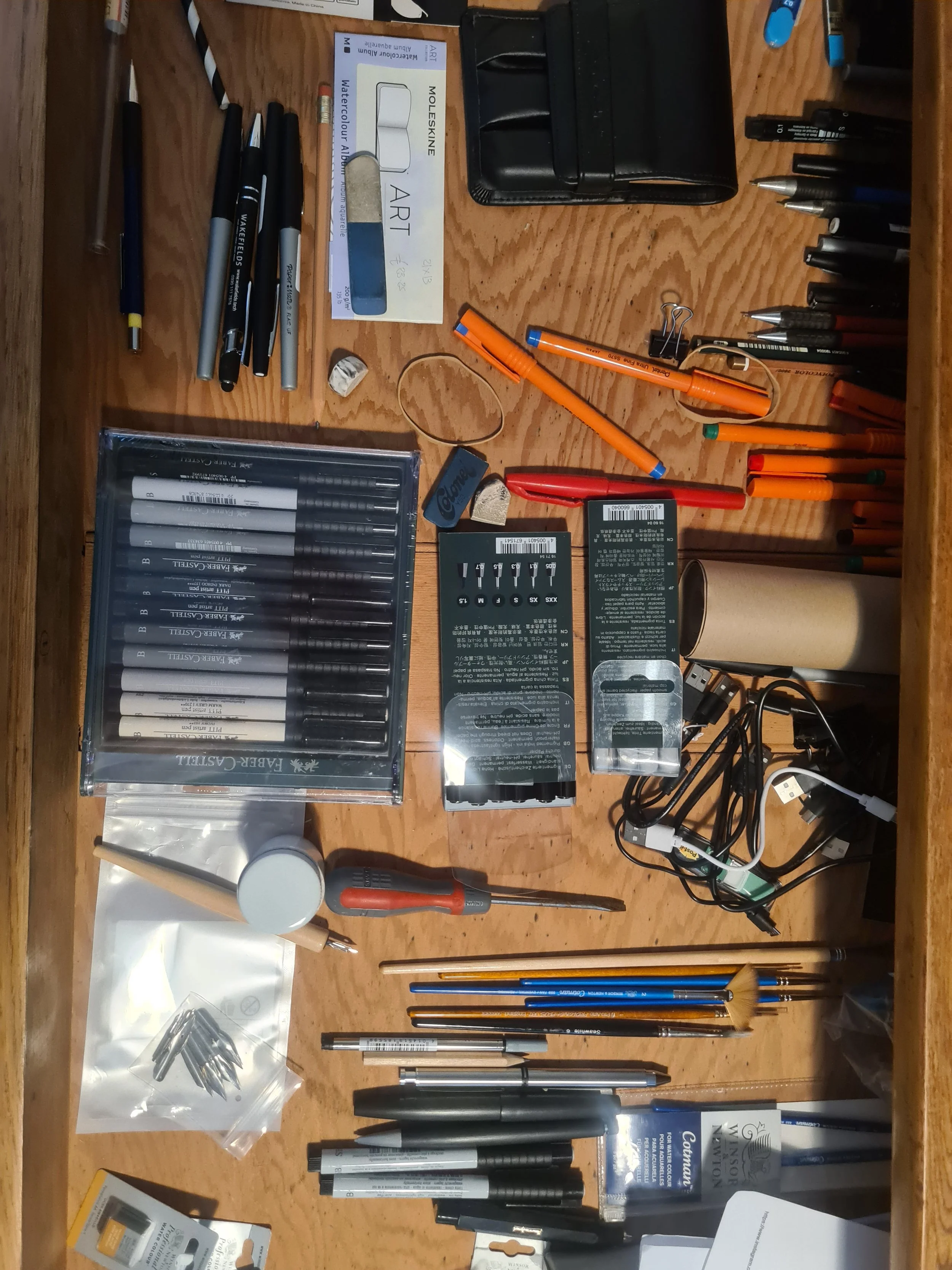 Assorted pens, markers, and art supplies on a wooden desk, with various small tools, batteries, and cables scattered around.