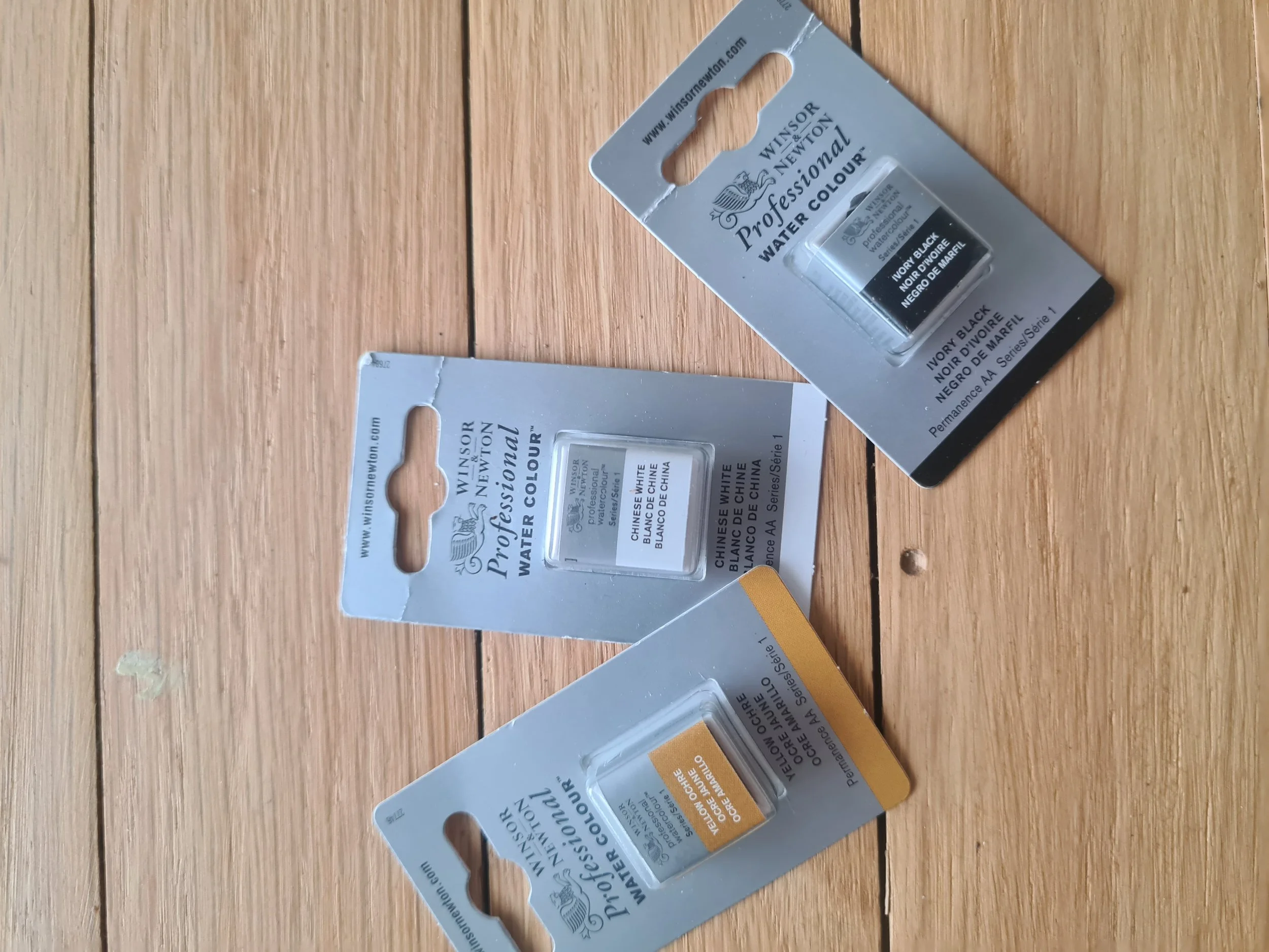 Three small tubes of Winsor & Newton professional watercolors in white, black, and orange, on a wooden surface. The tubes are in cardboard packaging with clear plastic windows.