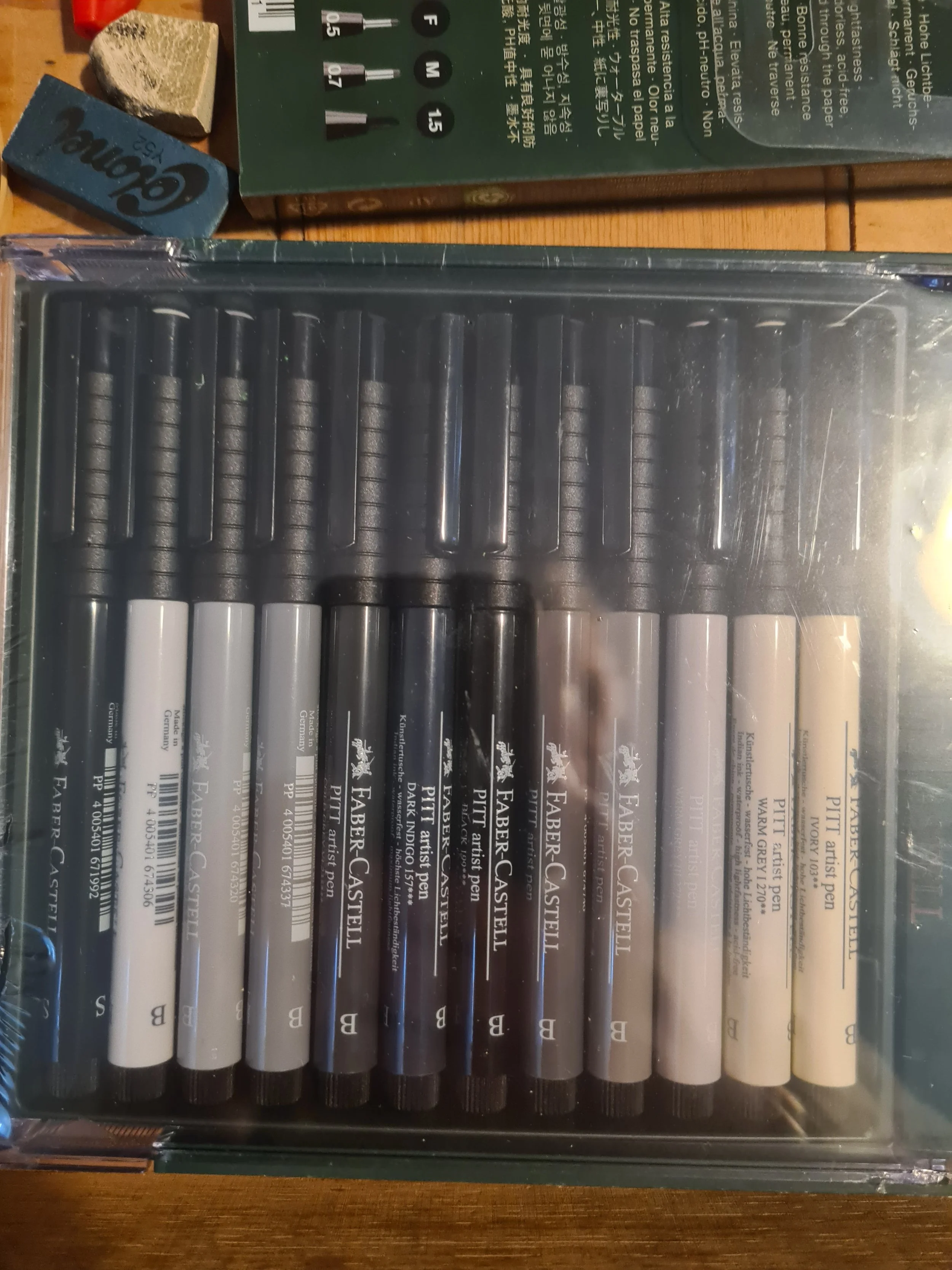 A box of Faber-Castell black, white, and beige ink pens in plastic packaging, arranged in a row inside a transparent case.