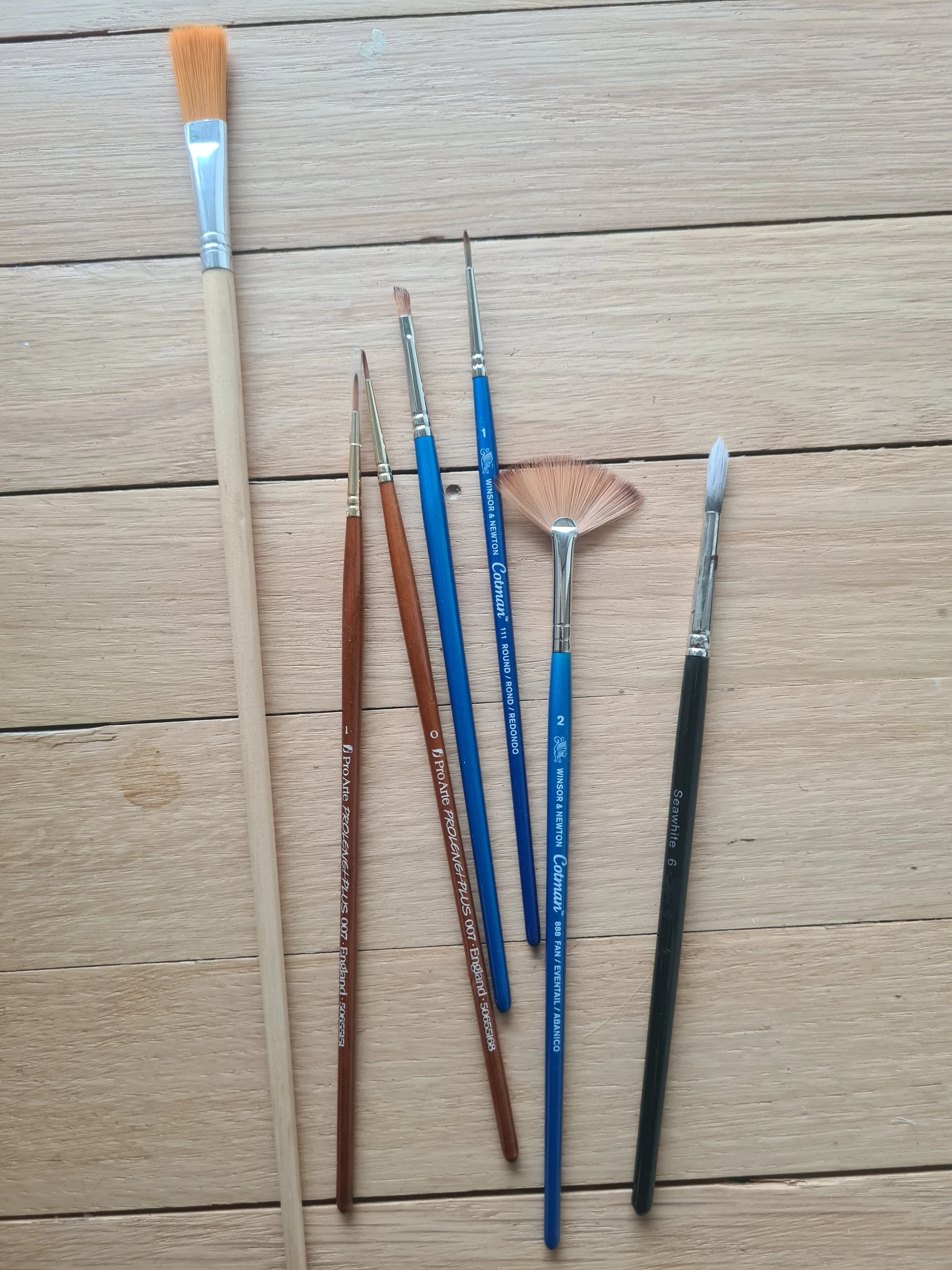 Set of seven paintbrushes arranged on a wooden surface, varying in size and shape.