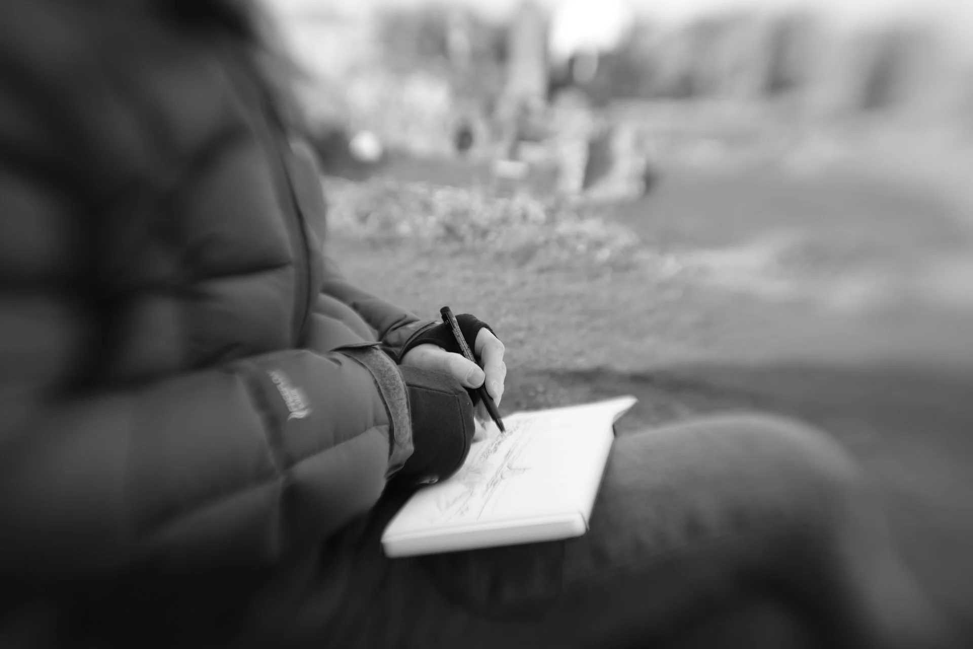 Person writing in a notebook outdoors, wearing gloves and a puffy jacket, with trees and rocks in the background.