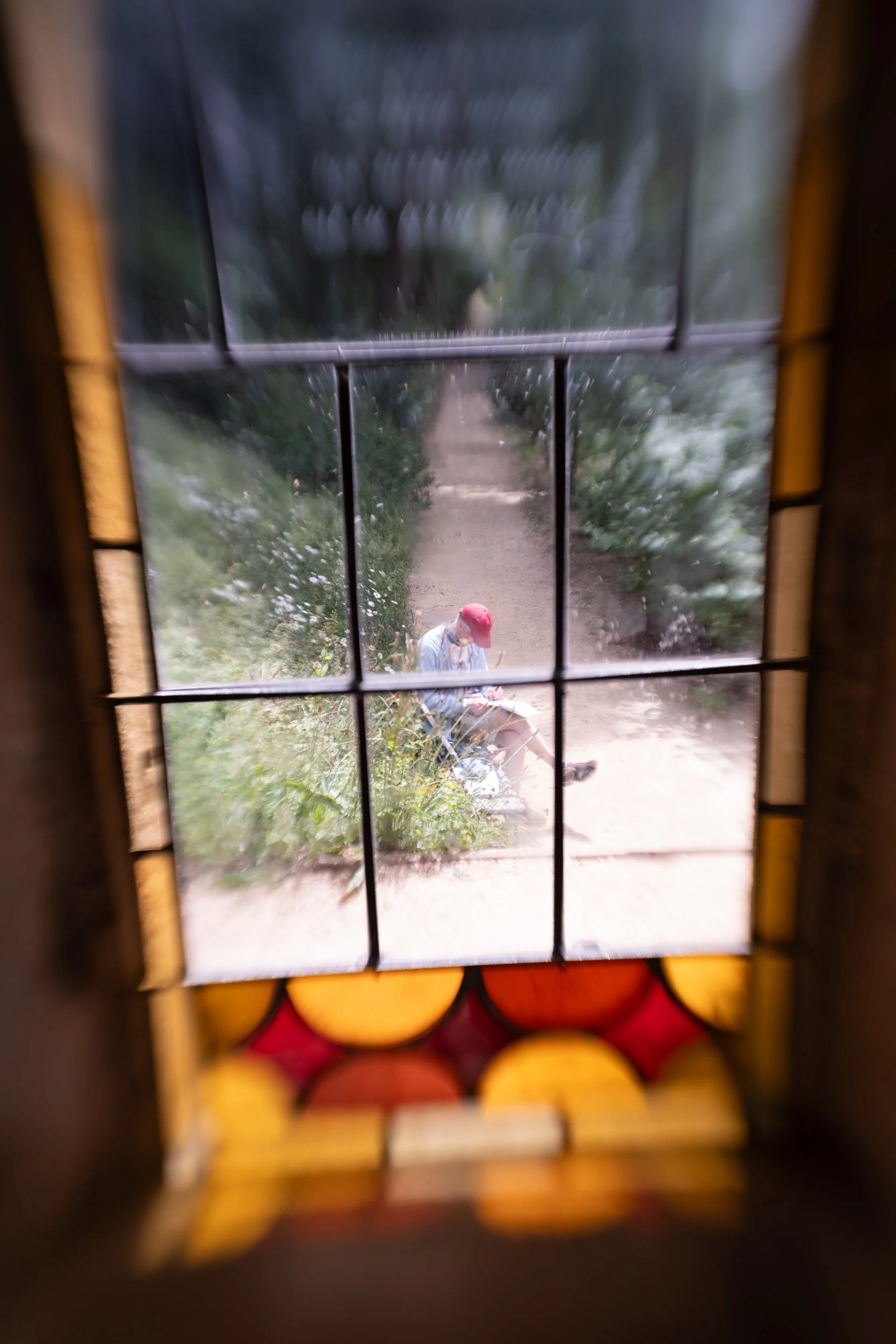 View through a stained glass window showing a person sitting on a bench reading, wearing a red hat and face mask, on a sidewalk with greenery.