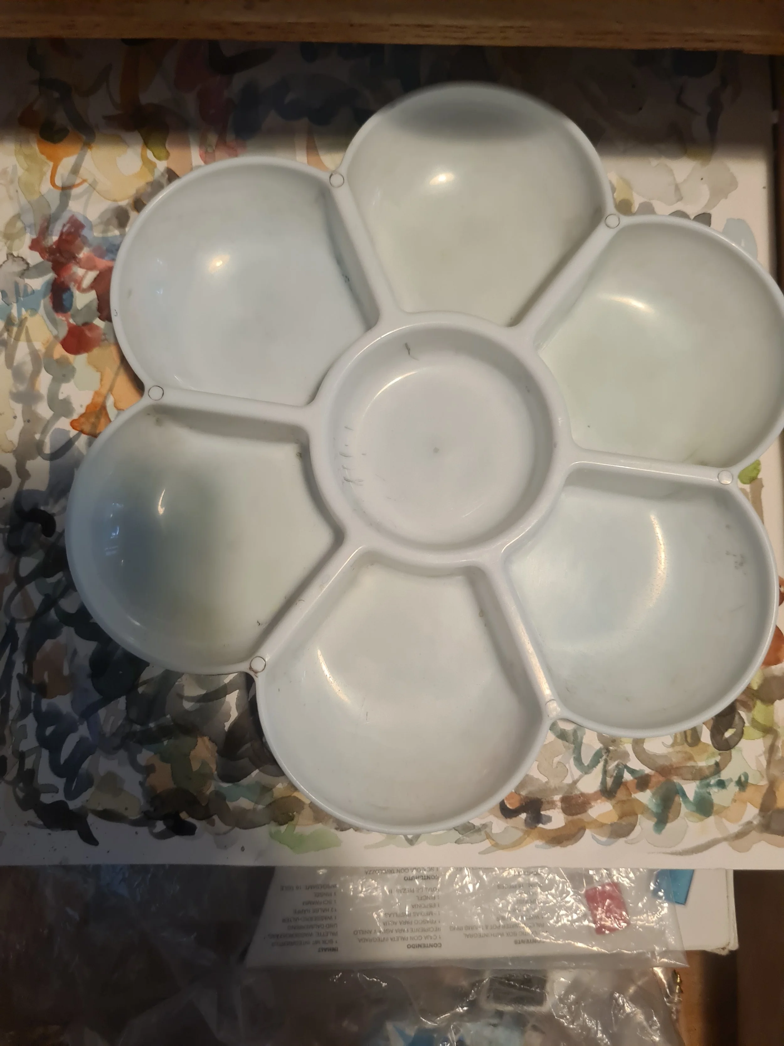 A white plastic flower-shaped paint palette with six petal sections and a central circular section, resting on a colorful, abstract background.