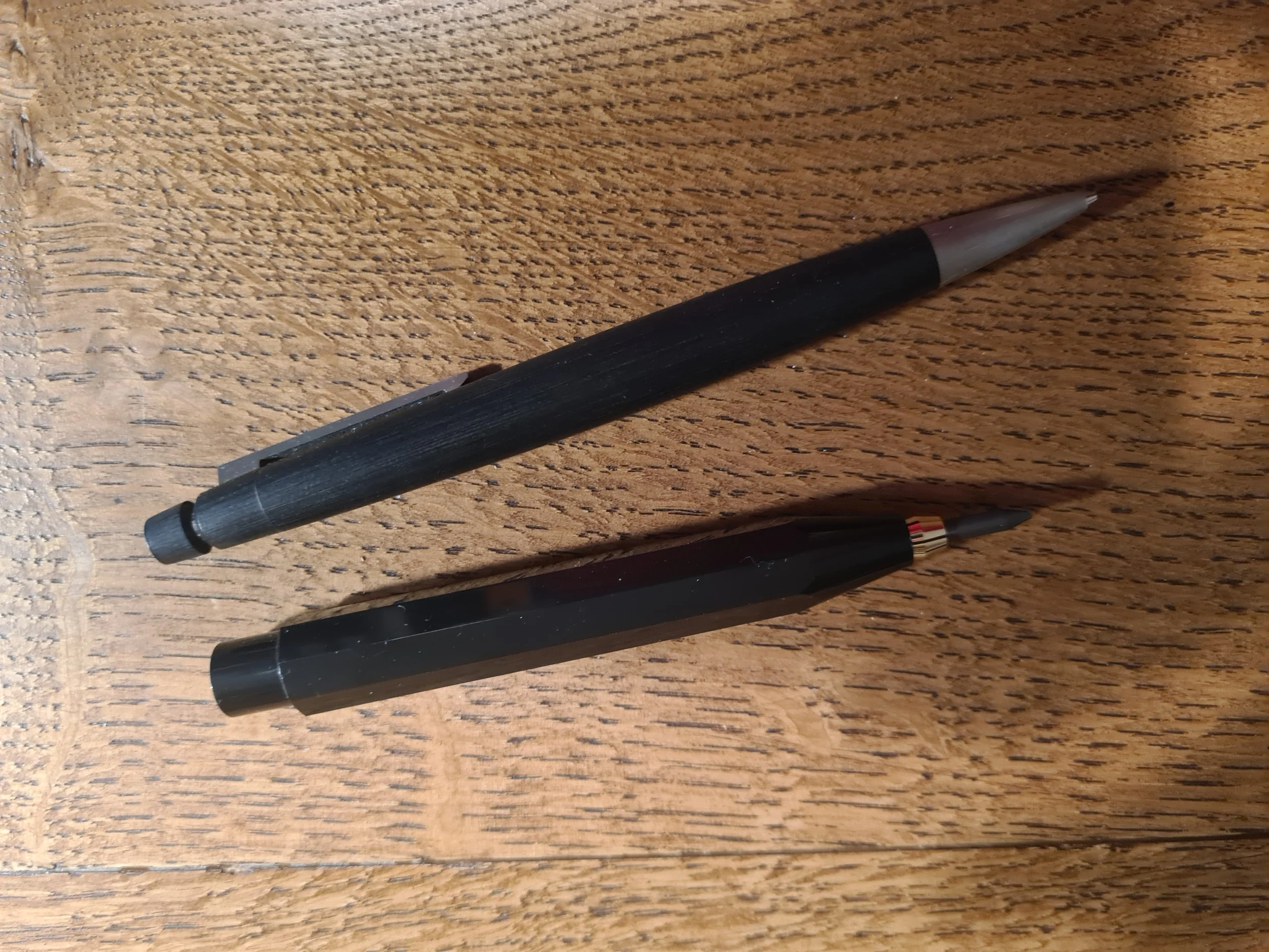 Two black mechanical pencils with erasers, one with a matte finish and one with a glossy finish, lying on a wooden surface.