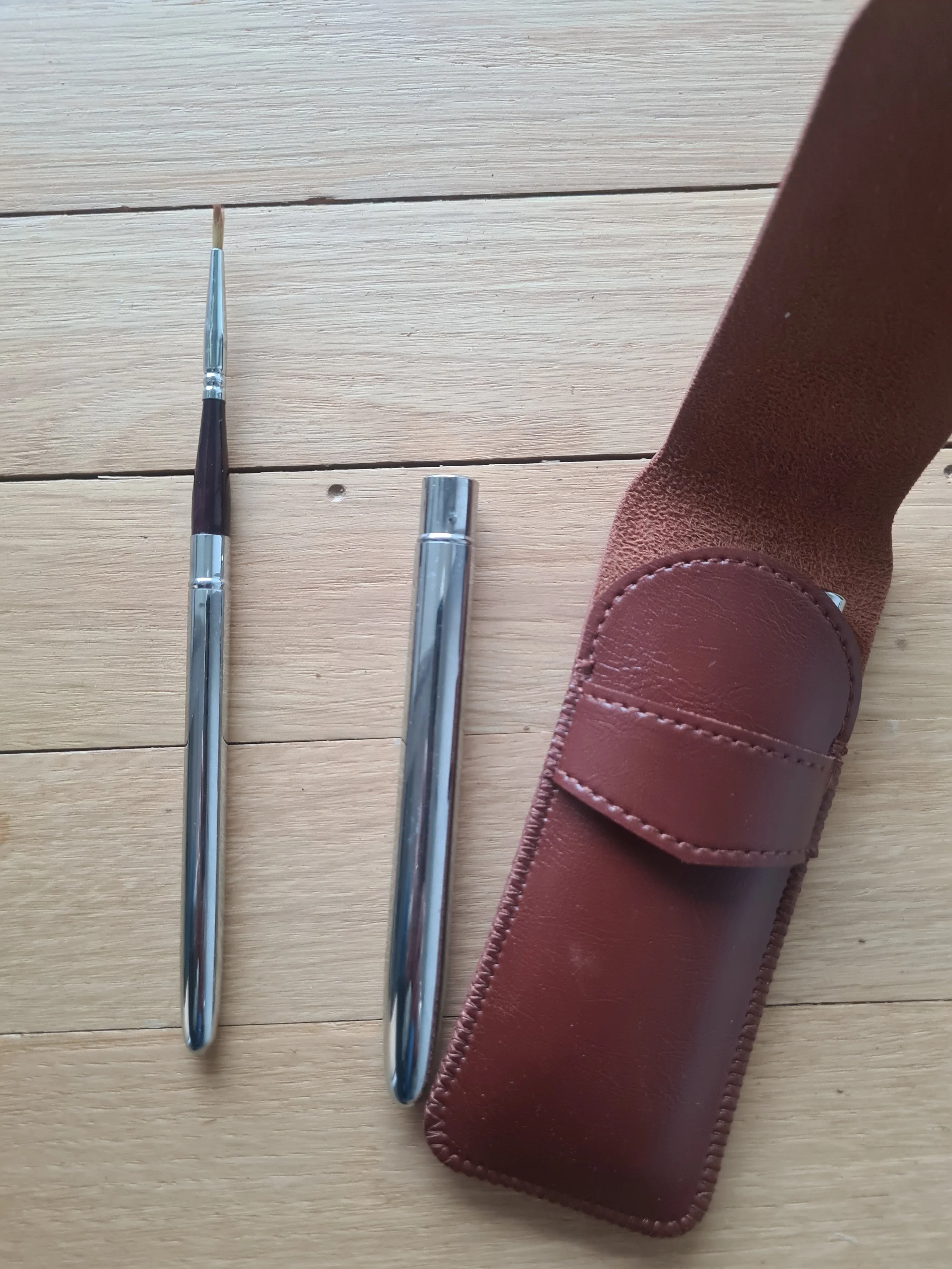 A pair of gardening tools, a small paintbrush and a tool with a curved metal end, lying on a wooden surface next to a brown leather sheath or holder.