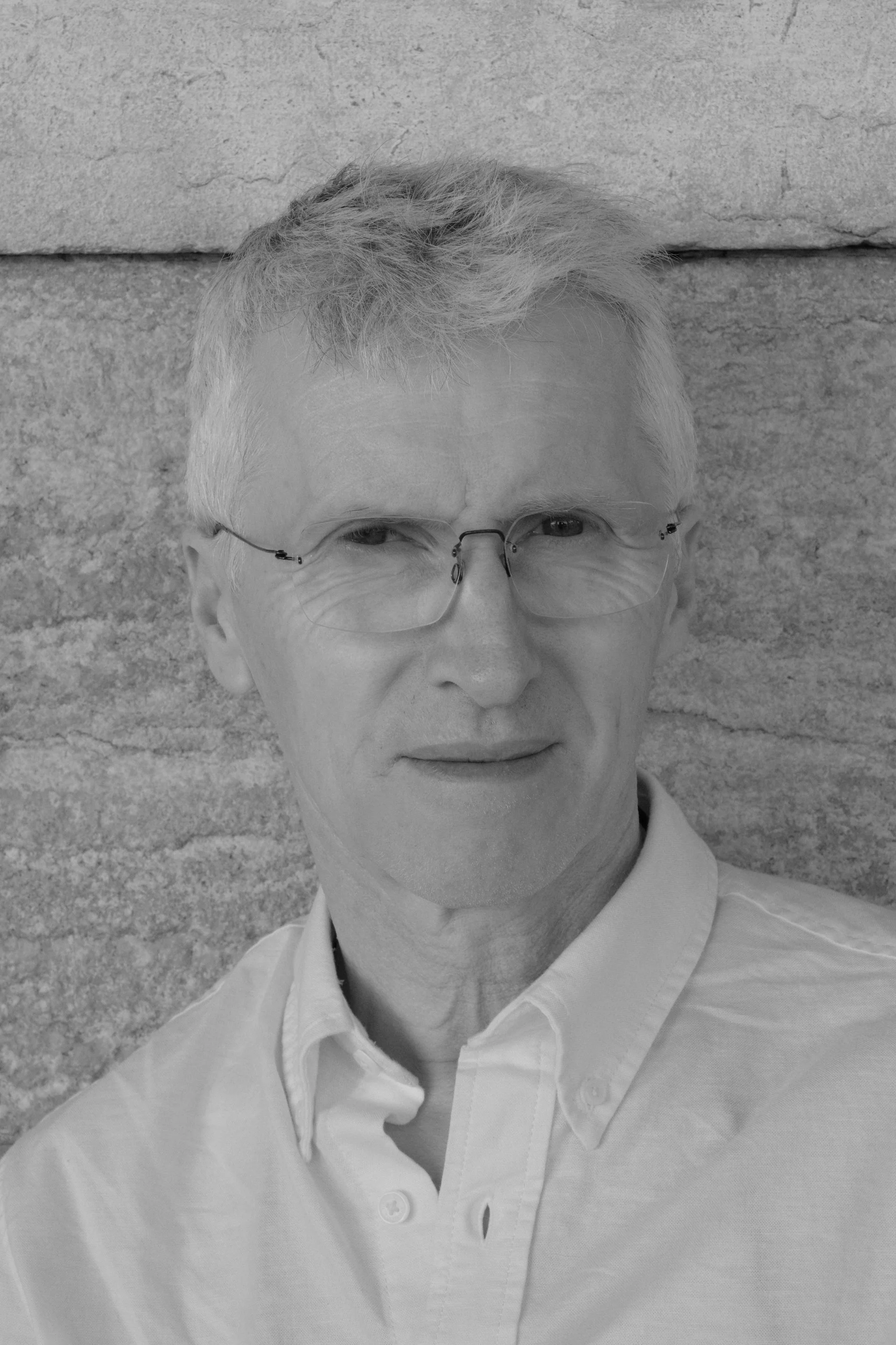 A black and white portrait of an older man with light hair, wearing glasses and a button-up shirt, standing against a textured stone wall.