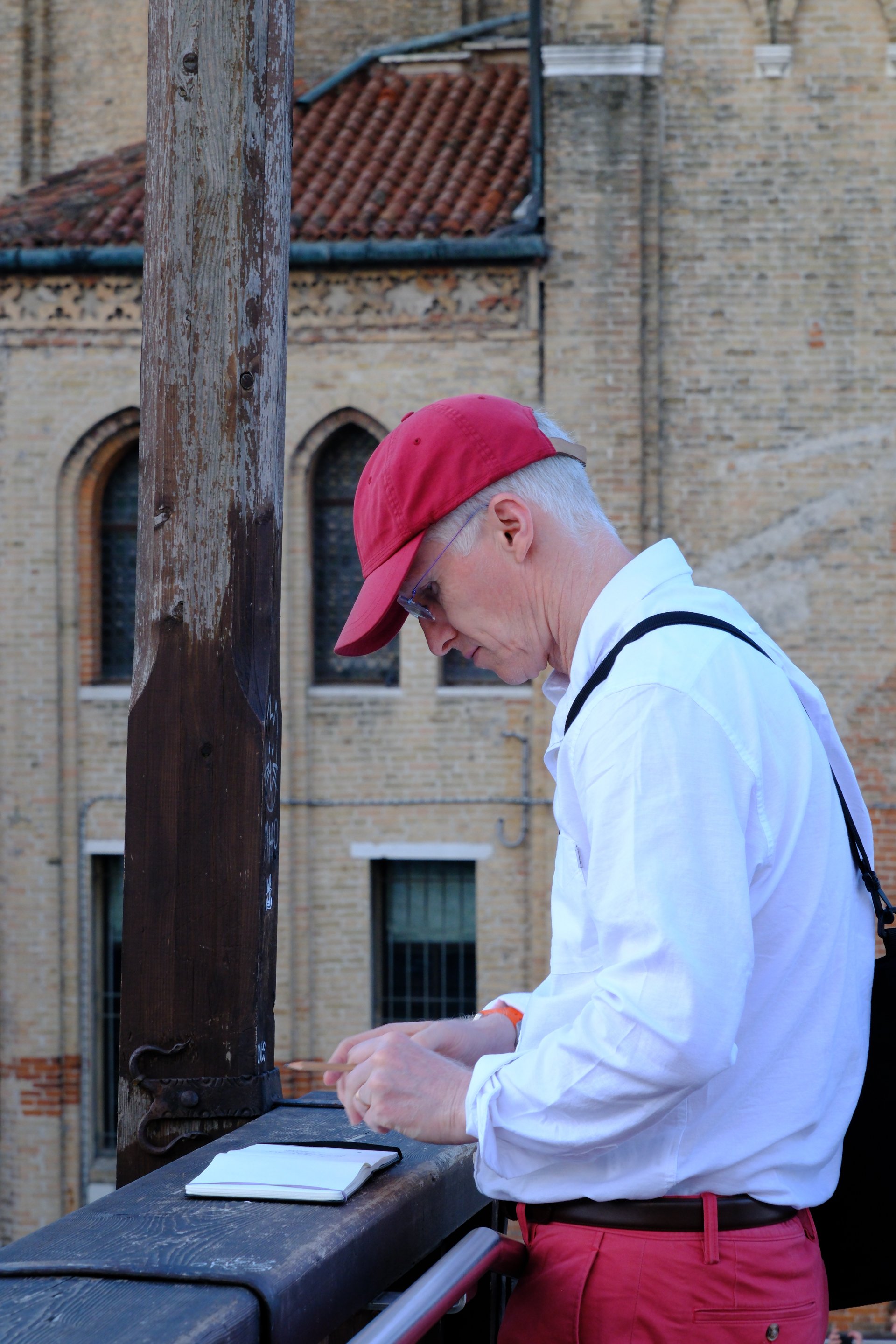 An older man wearing a red cap, white shirt, and red pants, standing outside near a wooden post and a building with brick walls and arched windows, looking at his phone.