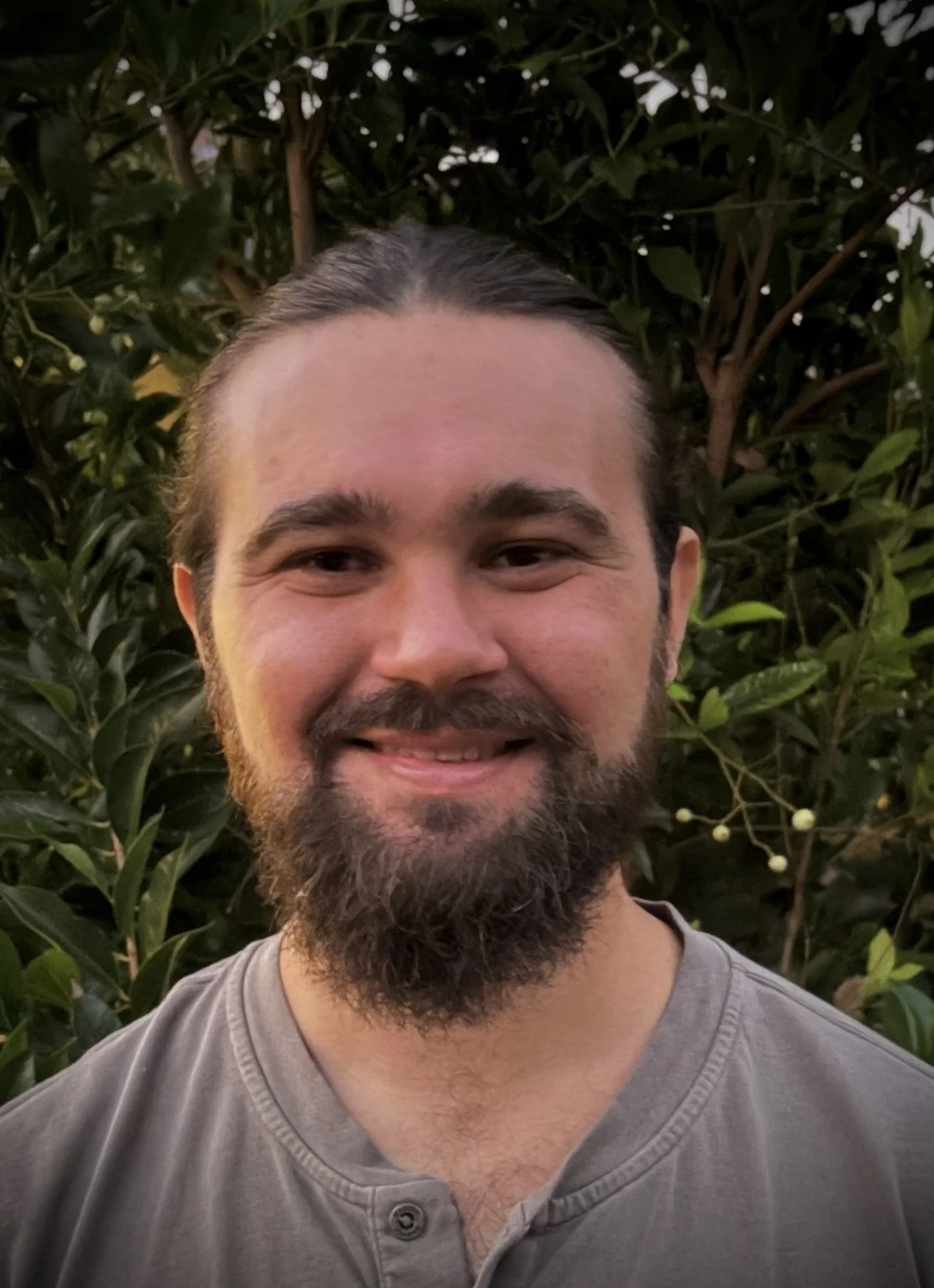 A smiling man with a beard and long hair pulled back, standing outdoors in front of green leafy bushes.