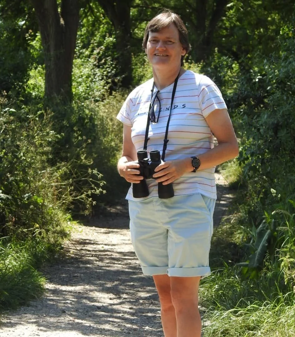 woman standing on a dirt path in a green forest, holding binoculars, wearing a striped t-shirt and shorts, with sunglasses hanging from her shirt and a watch on her wrist